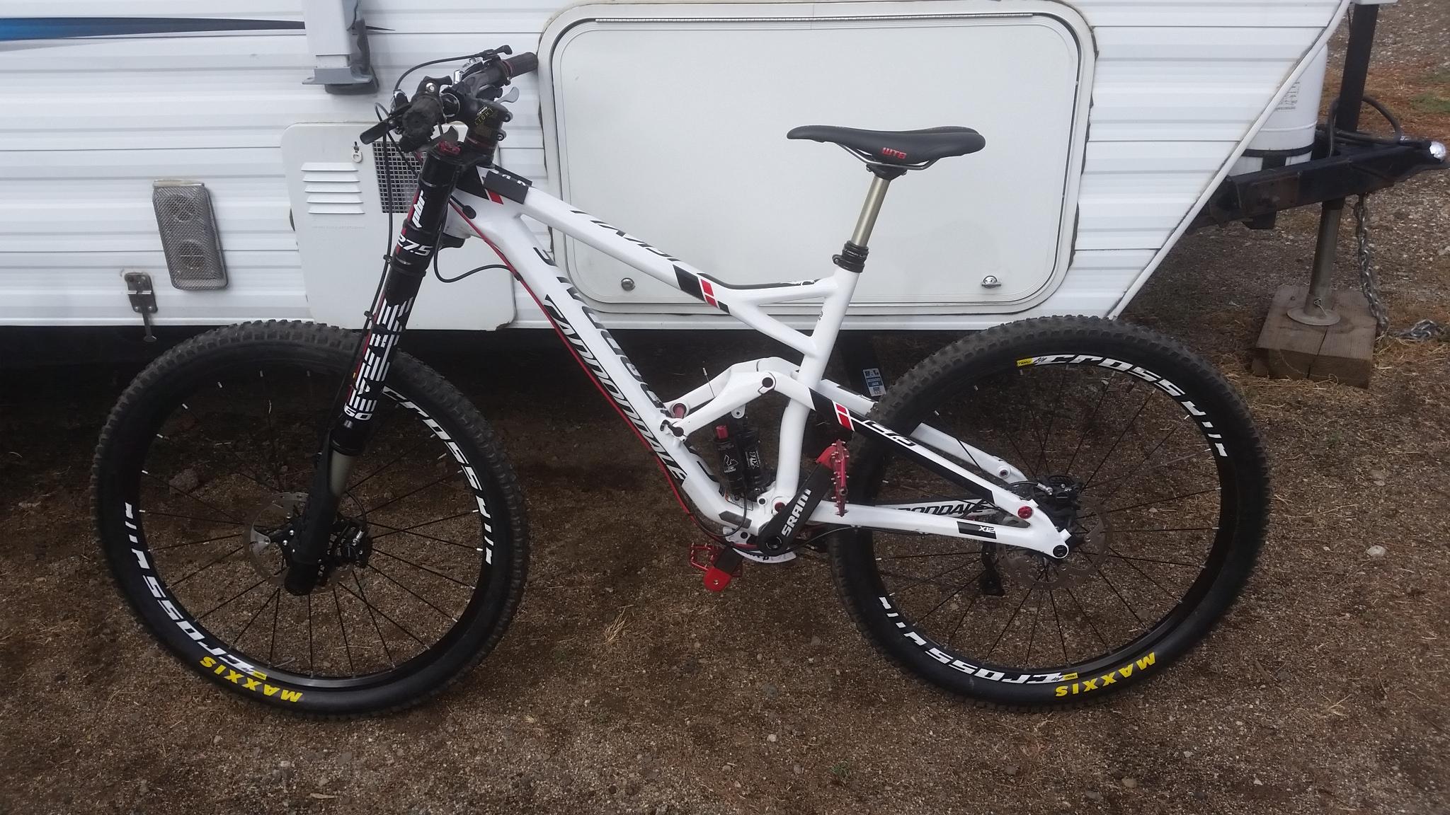 Cannondale Jekyll Carbon 2: A white mountain bike with red and black accents parked next to a parked camper trailer. The bike features wide tires, a dual suspension frame, and a prominent front fork. The ground is gravel, and the trailer has a horizontal white exterior with a grayish trim.
