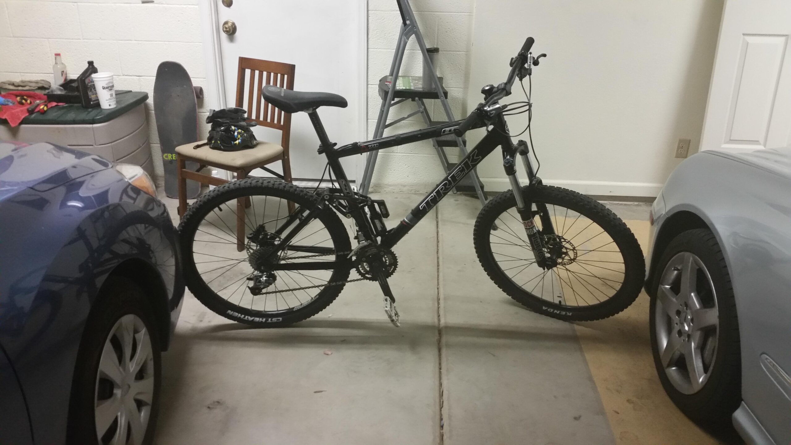 A mountain bike parked in a garage between two cars. The bike is black with a "Trek" logo and features thick tires for off-road riding. In the background, there is a chair and some storage containers along with a skateboard leaning against the wall.