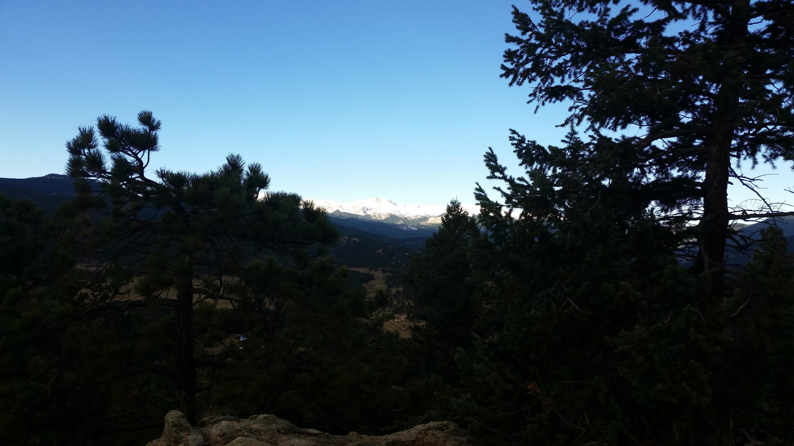 A scenic mountain view framed by evergreen trees, showcasing a clear blue sky and snow-capped peaks in the distance. The landscape includes a valley below, surrounded by hills and forests. 3 Sisters / Alderfer mountain bike trail.