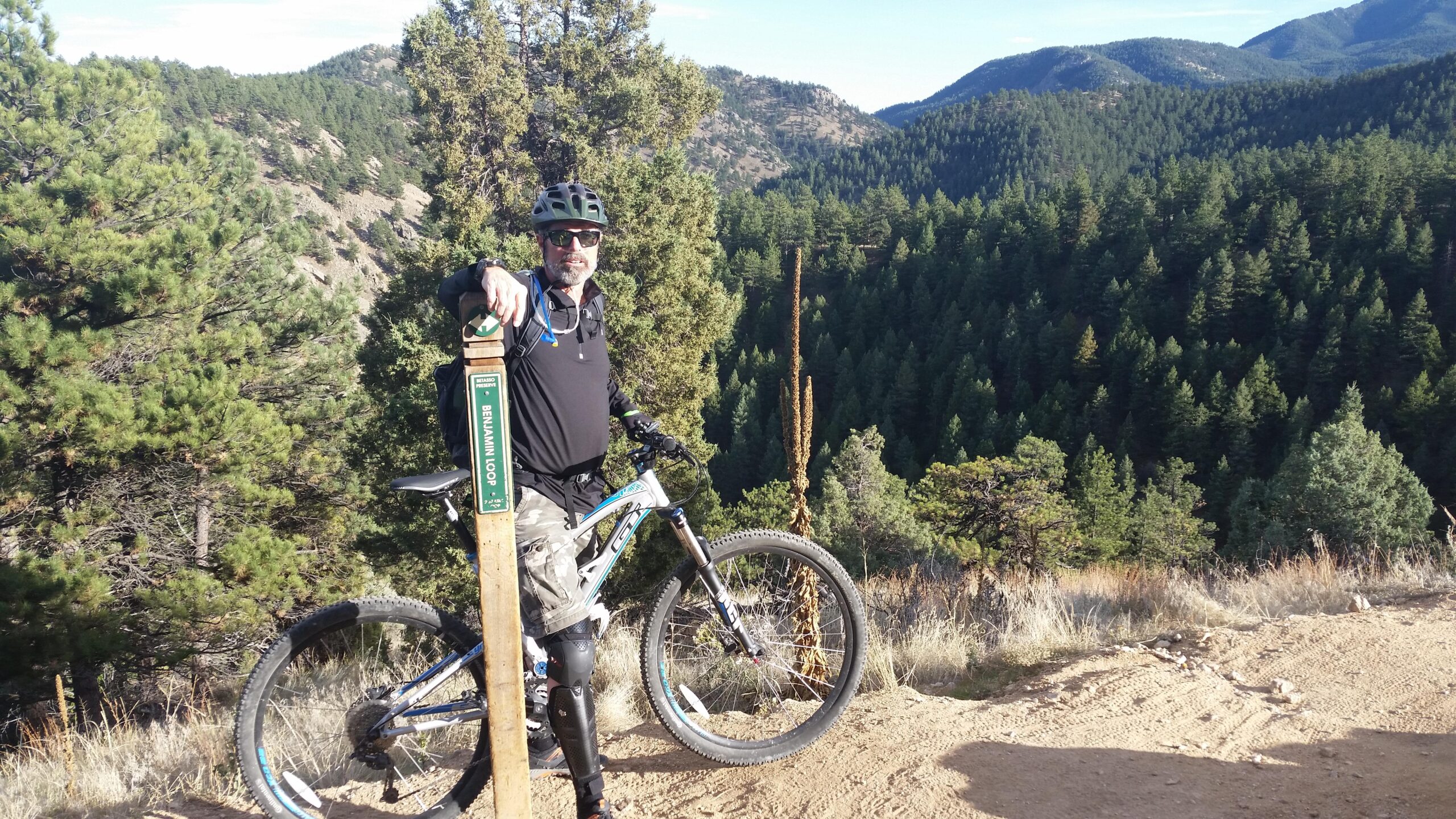 A mountain biker standing next to a trail sign labeled "Benjamin Woop" in a lush, green forested area, with mountains visible in the background. The biker is wearing a helmet and sunglasses, leaning against his bike, and the landscape features pine trees and rocky terrain under a clear sky. Betasso Preserve mountain bike trail.