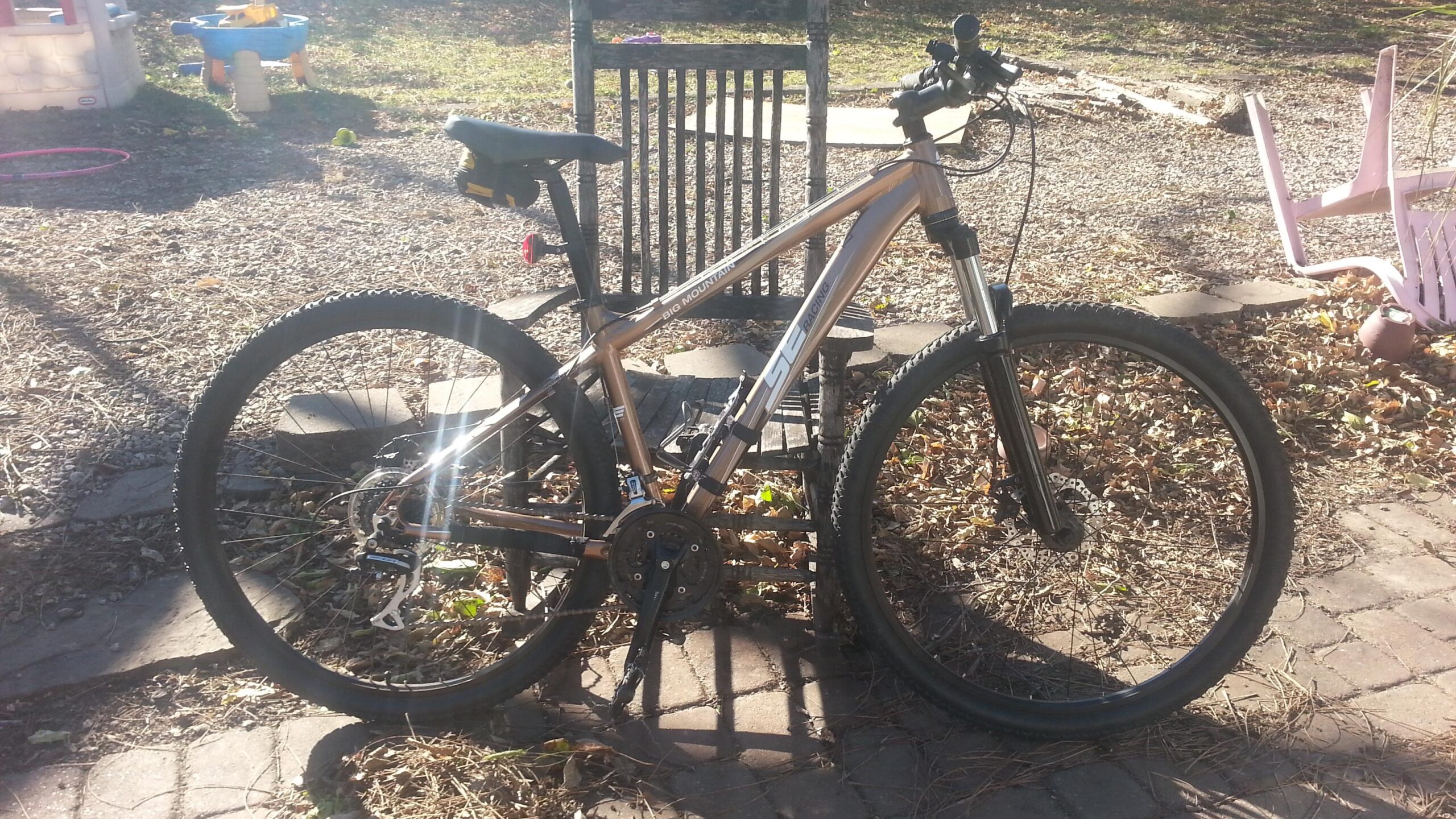 A mountain bike with a metallic gold frame is leaning against a wooden rocking chair in a yard. The ground is covered with leaves and small stones, and there are signs of play equipment in the background, including a plastic playhouse and a hula hoop. The scene is illuminated by sunlight, creating a bright and outdoor feel.