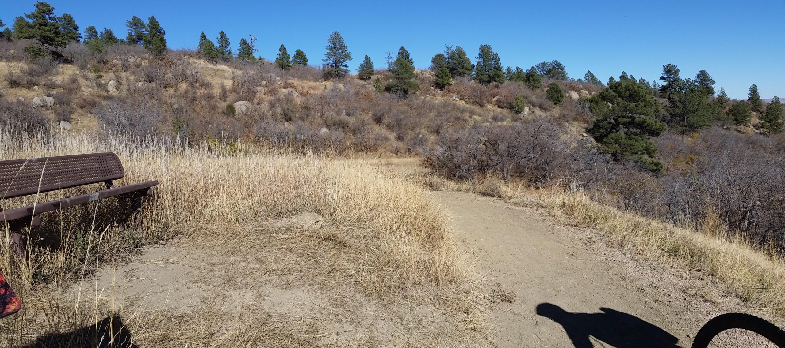 A scenic view of a dirt path winding through dry grass and shrubs, with a bench visible on the left. The background features hills covered in sparse vegetation and scattered trees under a clear blue sky. Cheyenne Mountain State Park mountain bike trail.