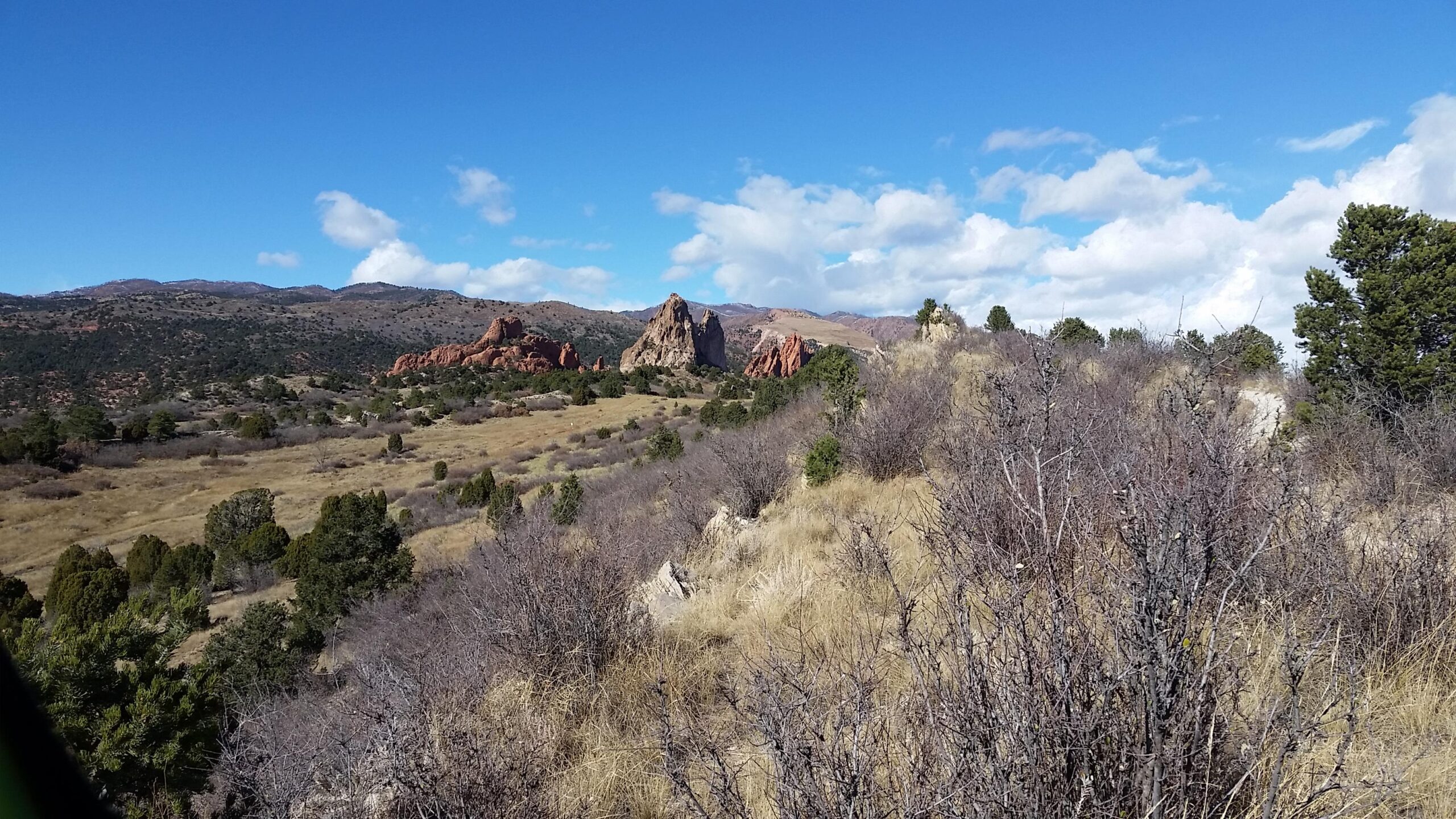 A scenic landscape featuring rolling hills, sparse vegetation, and distinctive rock formations against a backdrop of blue skies with scattered clouds. The foreground includes dry grass and bushes, while the background showcases rugged terrain and distant mountains. Garden of the Gods: Ute Trail mountain bike trail.