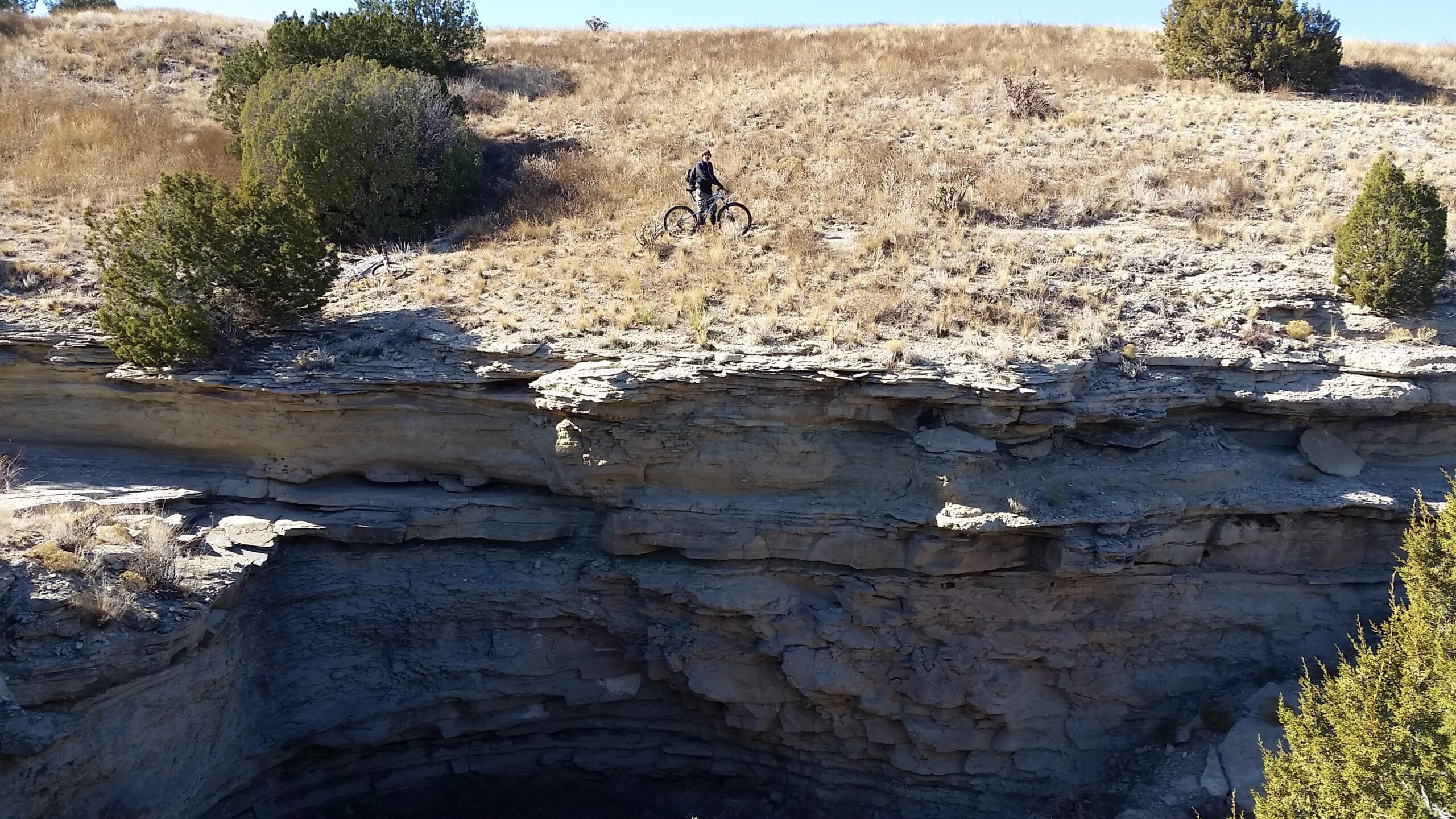 A mountain biker on a rocky ledge overlooking a deep geological depression, surrounded by dry grass and sparse vegetation under a clear blue sky. South Shore Lake Pueblo mountain bike trail.