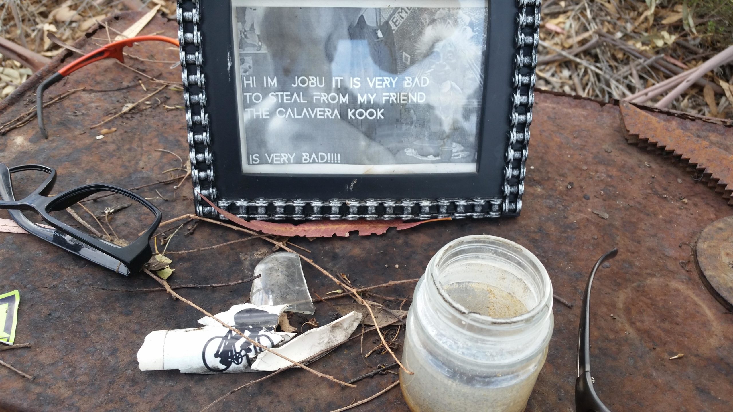 A framed note resting on a rusted metal surface, surrounded by leaves and debris. The note reads, "HI I'M JOBU. IT IS VERY BAD TO STEAL FROM MY FRIEND THE CALAVERA KOOK IS VERY BAD!!!!" Nearby, there's a pair of black glasses, a broken piece of glass, and a small jar filled with sand. Lake Calavera mountain bike trail.