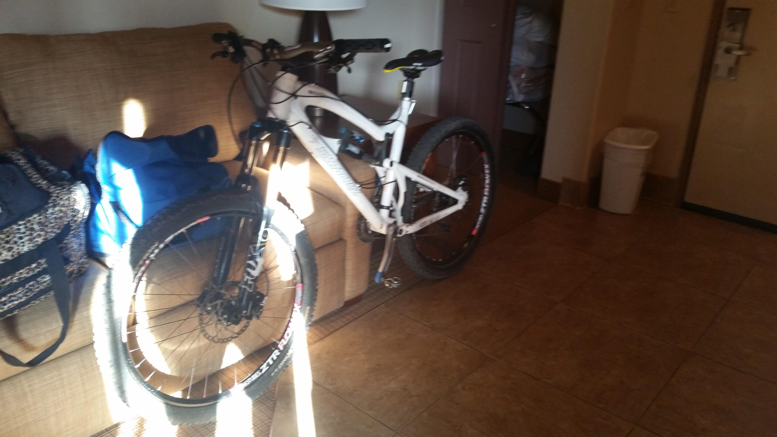 Santa Cruz Nomad: A mountain bike parked next to a couch in a room, with a blue bag and a black backpack nearby. Sunlight casts shadows on the floor and bike, highlighting the tiled flooring. A trash can is visible in the corner.