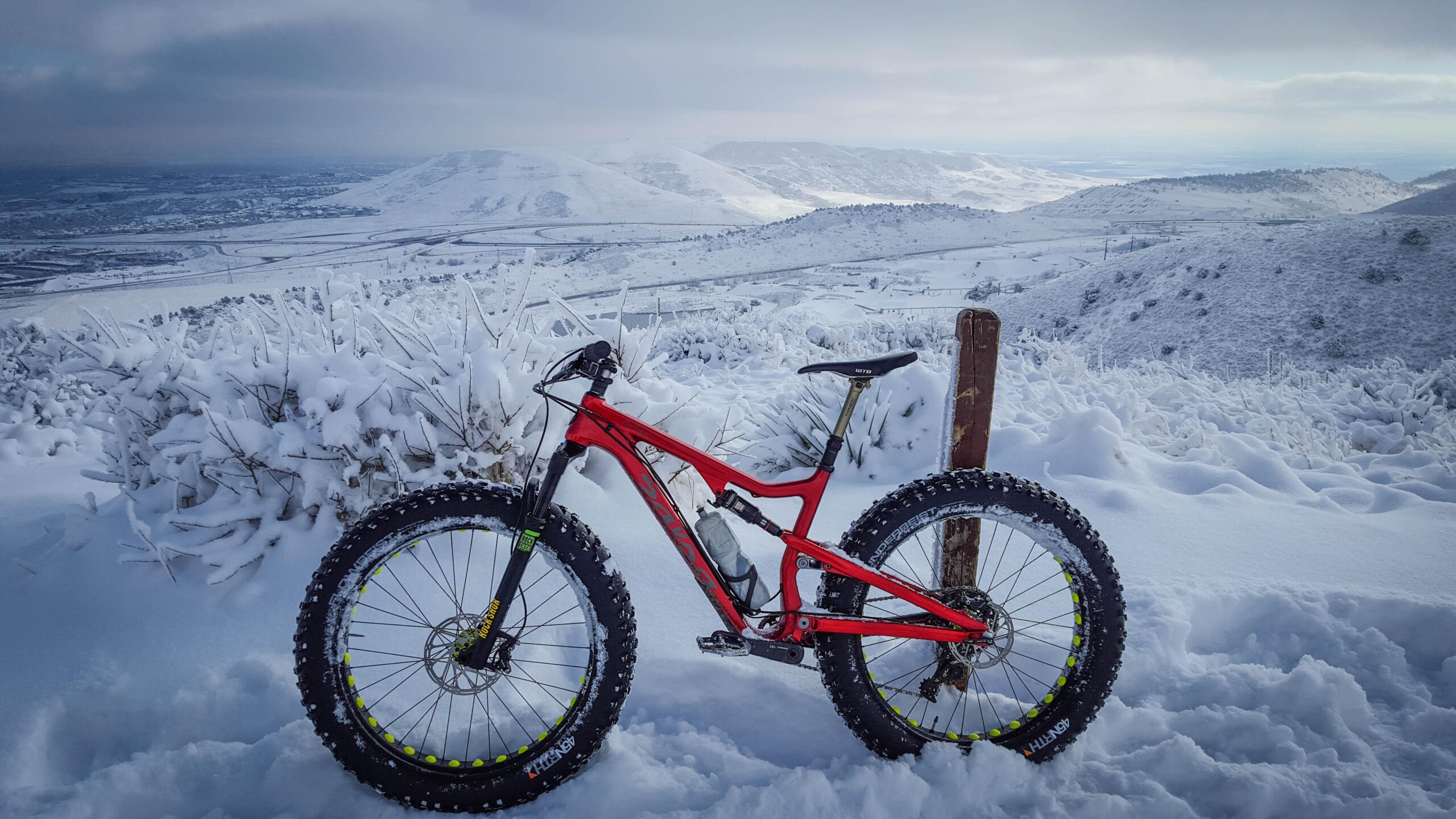 A bright red fat tire mountain bike parked in deep snow, with a snowy landscape and distant mountains in the background. The scene conveys a winter outdoor adventure setting. Apex Park mountain bike trail.