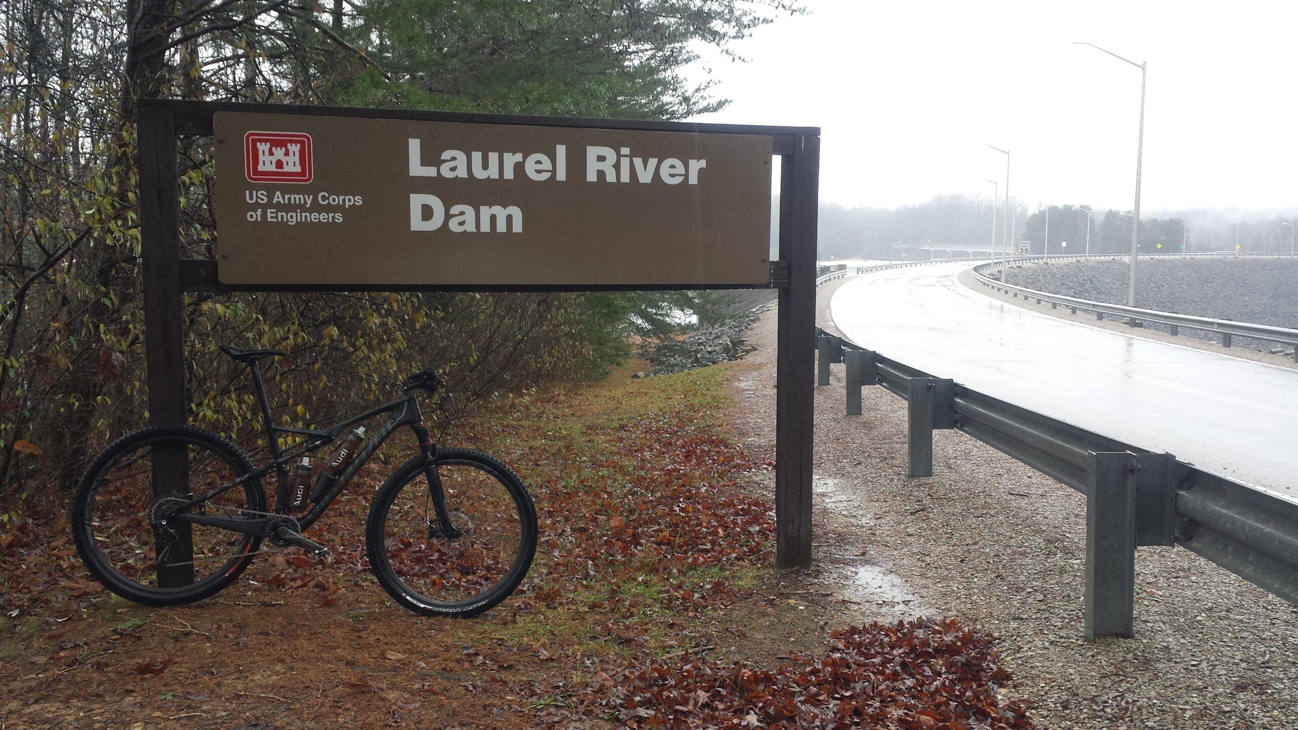 A brown sign marking the Laurel River Dam, with the U.S. Army Corps of Engineers logo, alongside a wet road. A bicycle is leaning against the sign, surrounded by fallen leaves and sparse vegetation on a rainy day. Sheltowee Trace - Laurel Lake Trail mountain bike trail.