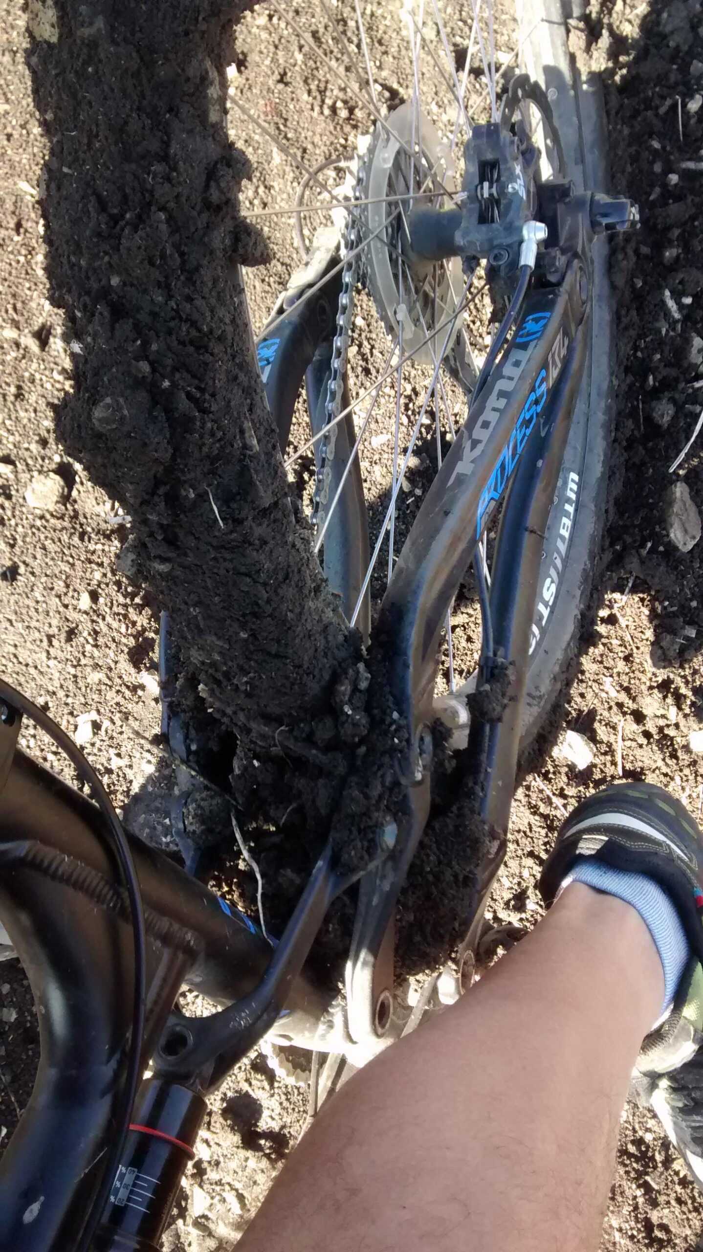 Kona Process 134 DL: A close-up view of a mountain bike with a heavily caked mud on its rear wheel and frame, showing the chain and brake components. A person's leg, wearing a sock and a shoe, is partially visible beside the bike on a dirt surface.