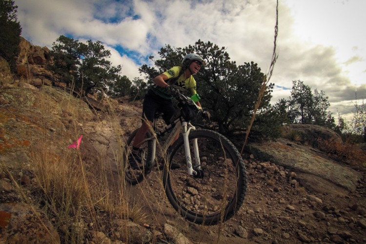 A mountain biker in a green and black jersey navigates a rocky trail, airborne with one wheel off the ground. Surrounding him are trees and rocky terrain under a partly cloudy sky, with a pink marker visible in the foreground.