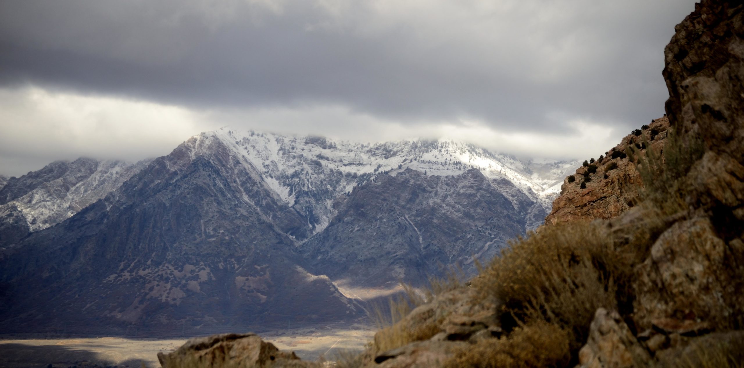 Majestic snow-capped mountains rise under a cloudy sky, with rocky terrain and sparse vegetation in the foreground. The scene captures a dramatic interplay of light and shadow across the rugged landscape. Bonneville Shoreline Trail - Ogden Section mountain bike trail.