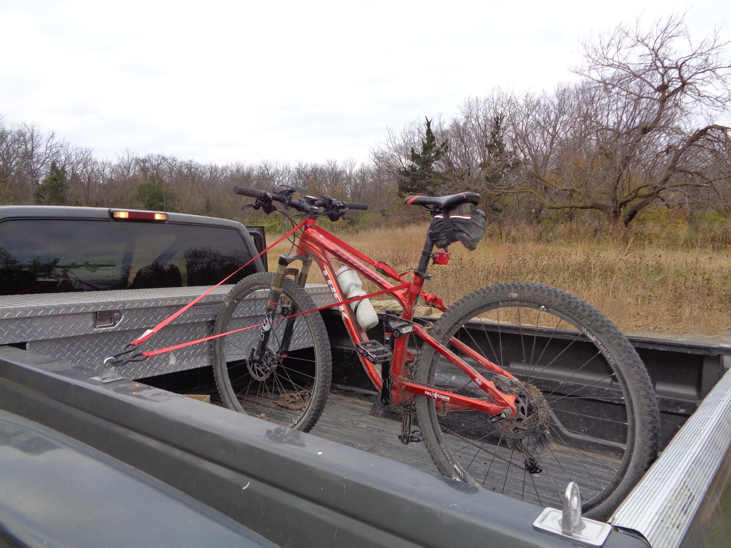 Mountain bike secured in the bed of a pickup truck, surrounded by a grassy area and bare trees under a cloudy sky. Skyline Park - Burnetts Mound Trail mountain bike trail.