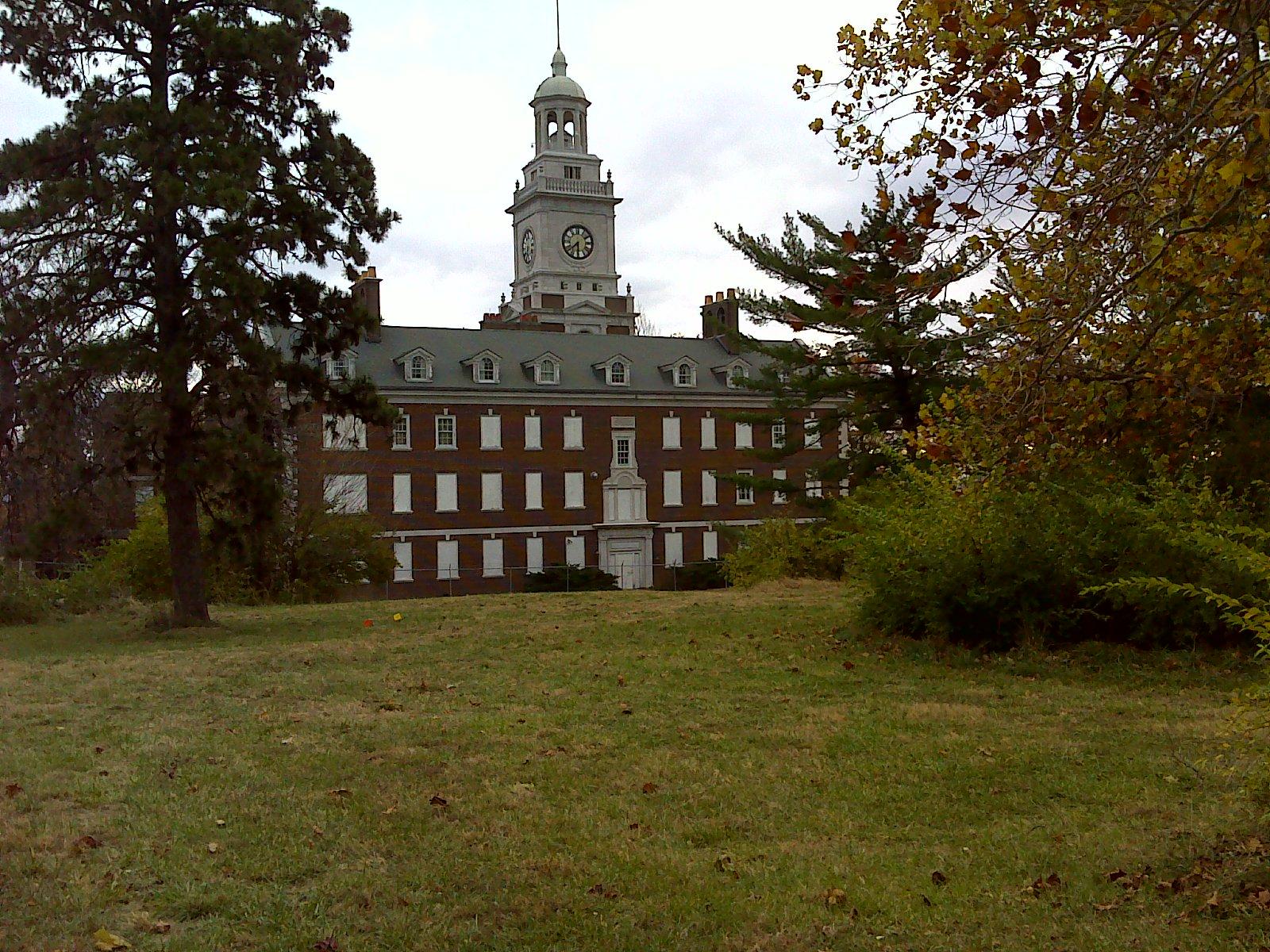 A historic building with a clock tower, surrounded by trees and grass, set against a cloudy sky. The architecture features red brick and white trim, with several windows and a front entrance. Maclennan Park mountain bike trail.
