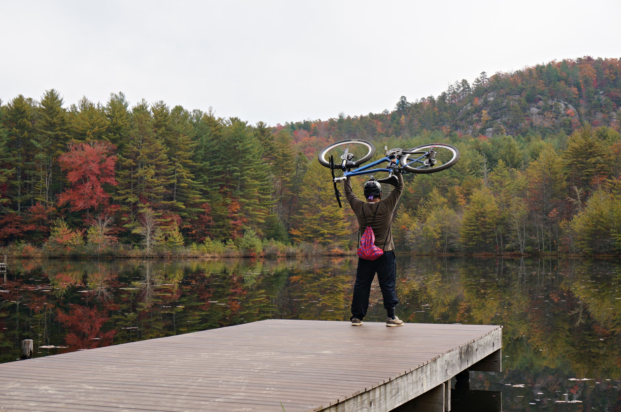 A person standing on a wooden dock by a calm lake, holding a bicycle overhead. The background features a colorful array of autumn foliage with red and green trees reflecting in the water. Cloudy skies are present above the scene. DuPont State Forest mountain bike trail.