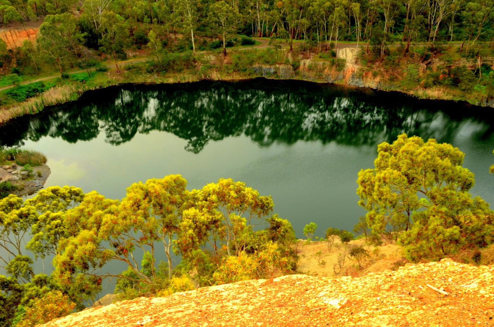 A serene view of a still, reflective pond surrounded by lush green trees and grassy banks, captured from a higher vantage point. The water mirrors the trees and landscape, creating a peaceful natural scene. Plenty Gorge mountain bike trail.
