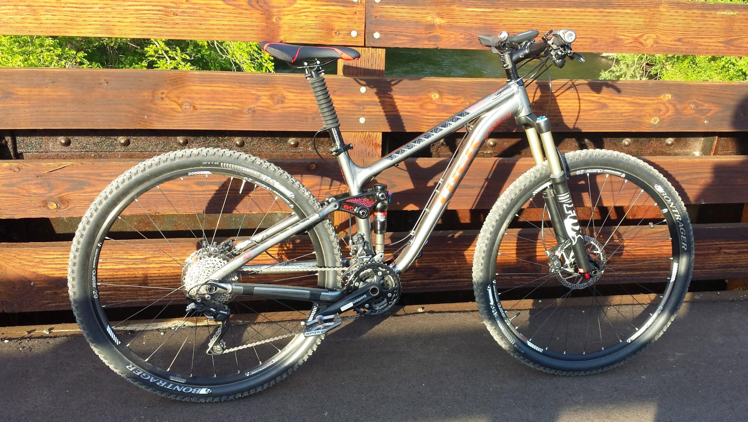 Trek Fuel EX 8 29: A modern mountain bike leaning against a wooden structure, showcasing its sleek silver frame, wide knobby tires, and front suspension fork. The bike is equipped with a red and black saddle, and the background features greenery peeking through the wooden slats.