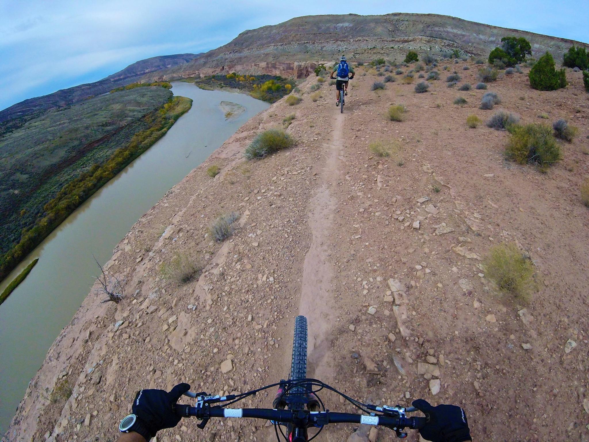 Mountain biker navigating a narrow dirt trail along a rocky cliff, with a river winding through a valley below. The landscape features sparse vegetation and rugged terrain under a cloudy sky. Kokopelli Area Trails mountain bike trail.