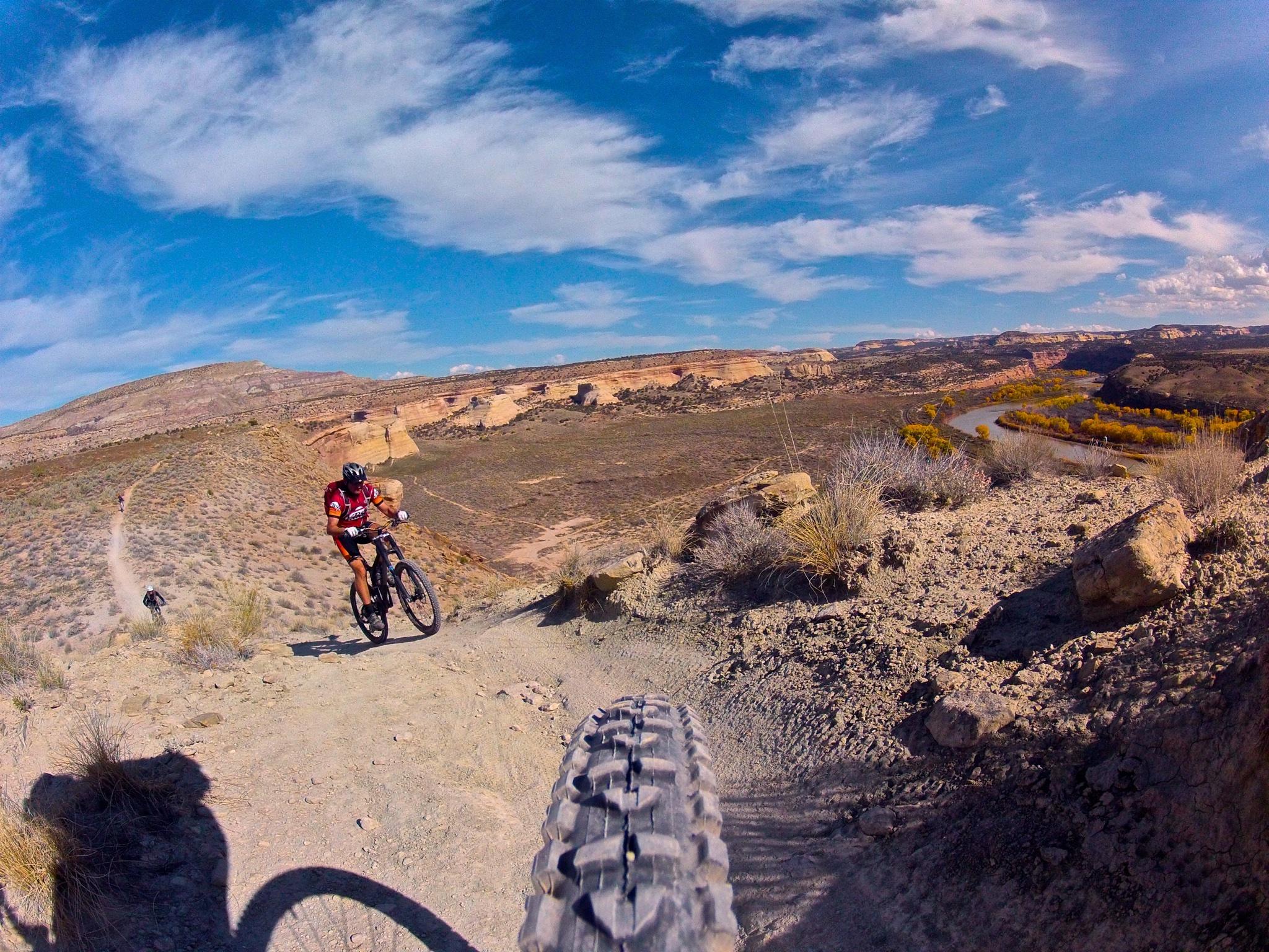 Mountain bikers navigating a rocky trail with stunning desert landscape and a river in the background under a clear blue sky. Western Rim mountain bike trail.