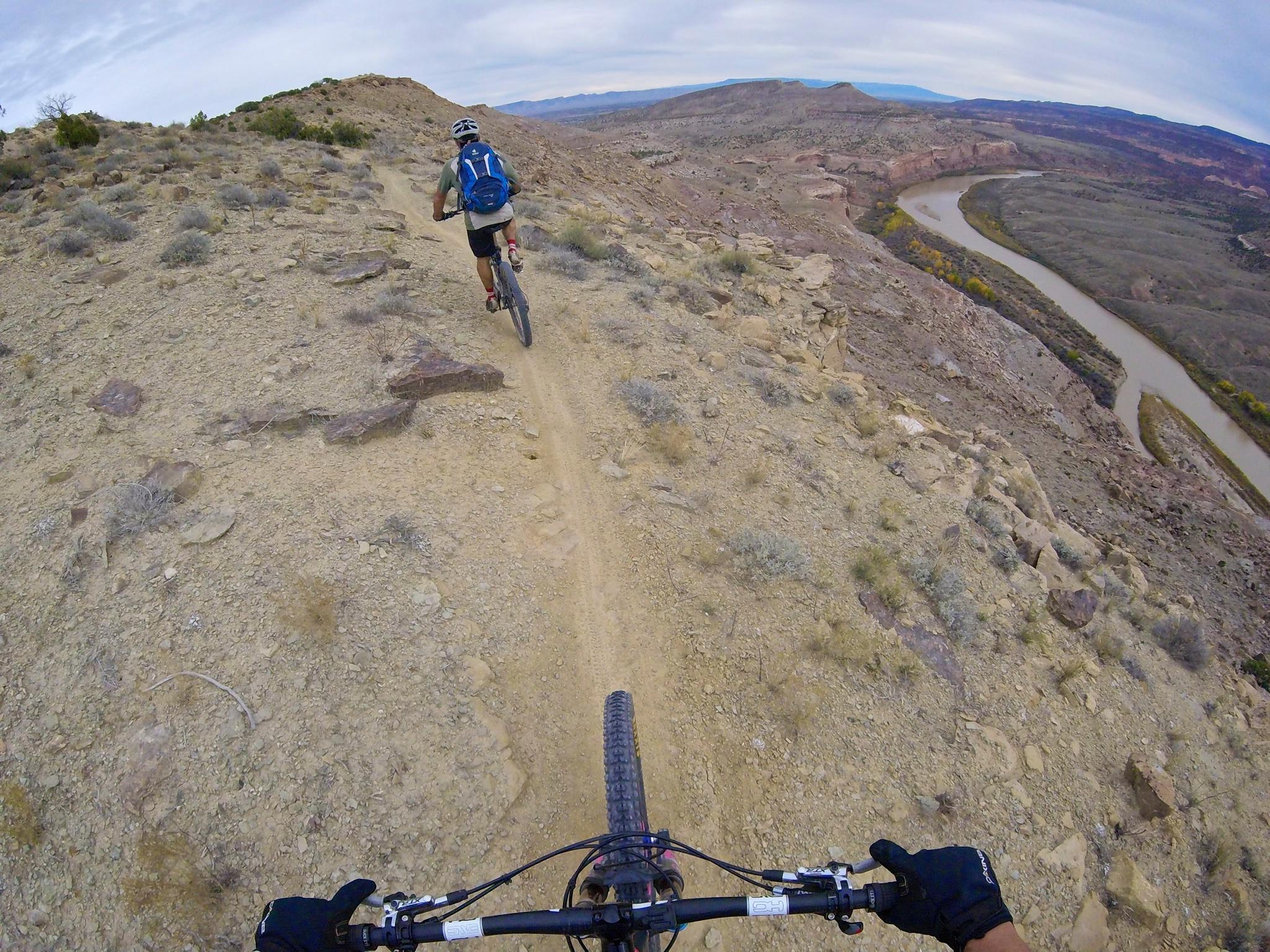 A mountain biker navigates a rugged trail on a hilltop with rocky terrain, overlooking a winding river and a vast landscape in the background. Kokopelli Area Trails mountain bike trail.