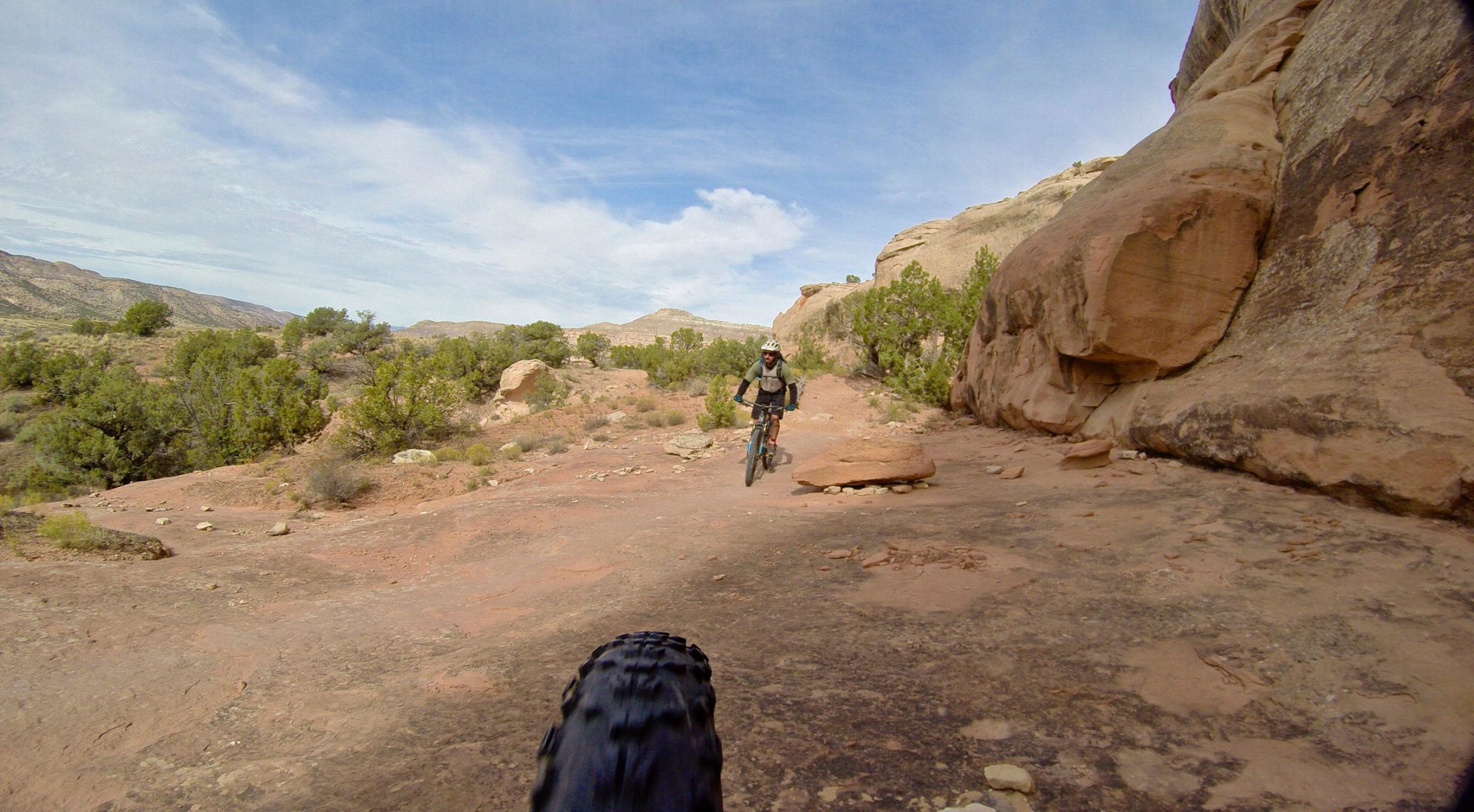 A cyclist riding on a rocky trail surrounded by desert vegetation and hills under a blue sky, with the front tire of the bike partially visible in the foreground. Kokopelli Area Trails mountain bike trail.