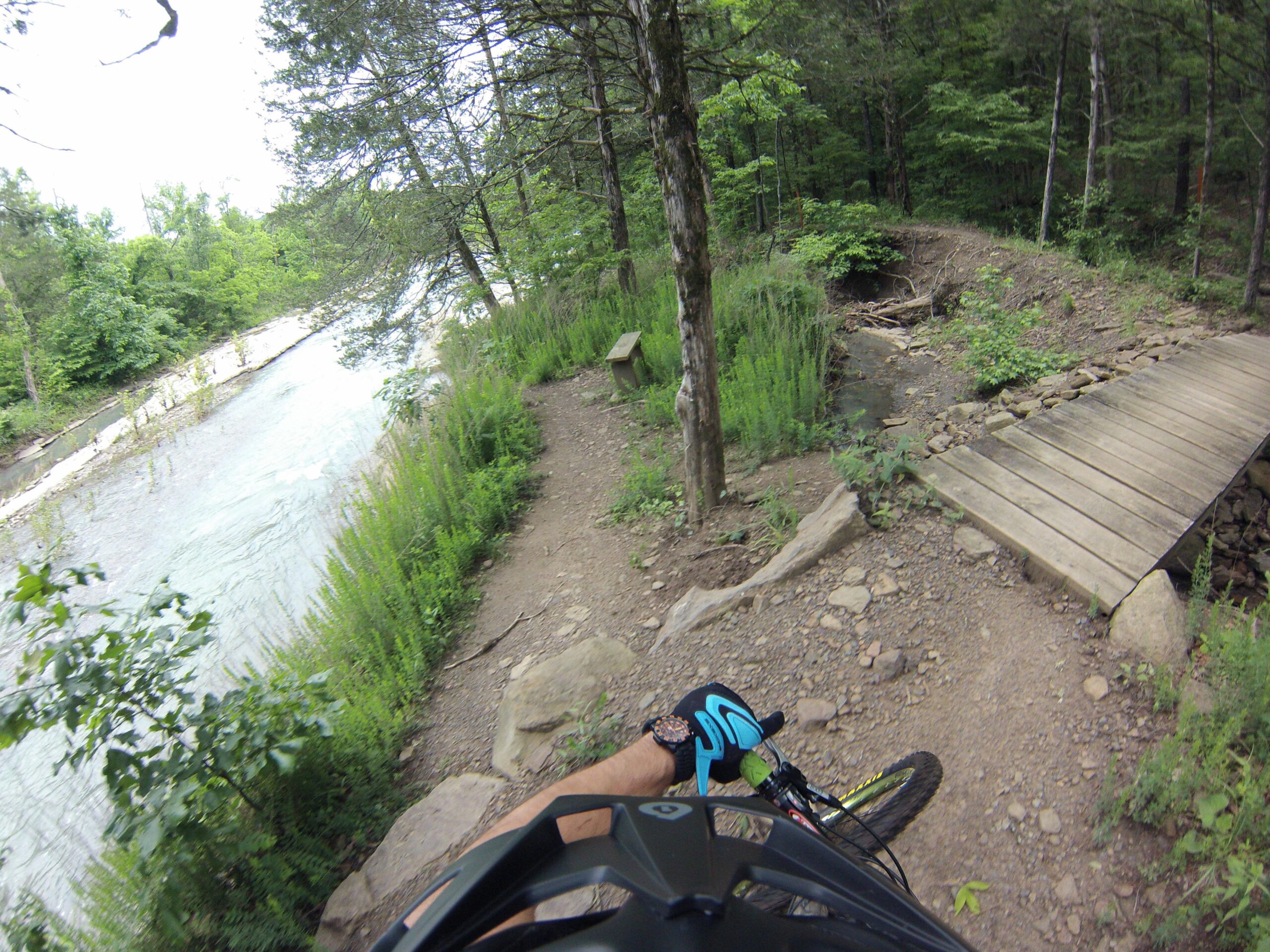 A cyclist's perspective of a rocky mountain biking trail alongside a flowing river, surrounded by lush green trees and vegetation. A wooden bridge crosses a small stream, with the trail winding ahead. Fossil Flats mountain bike trail.