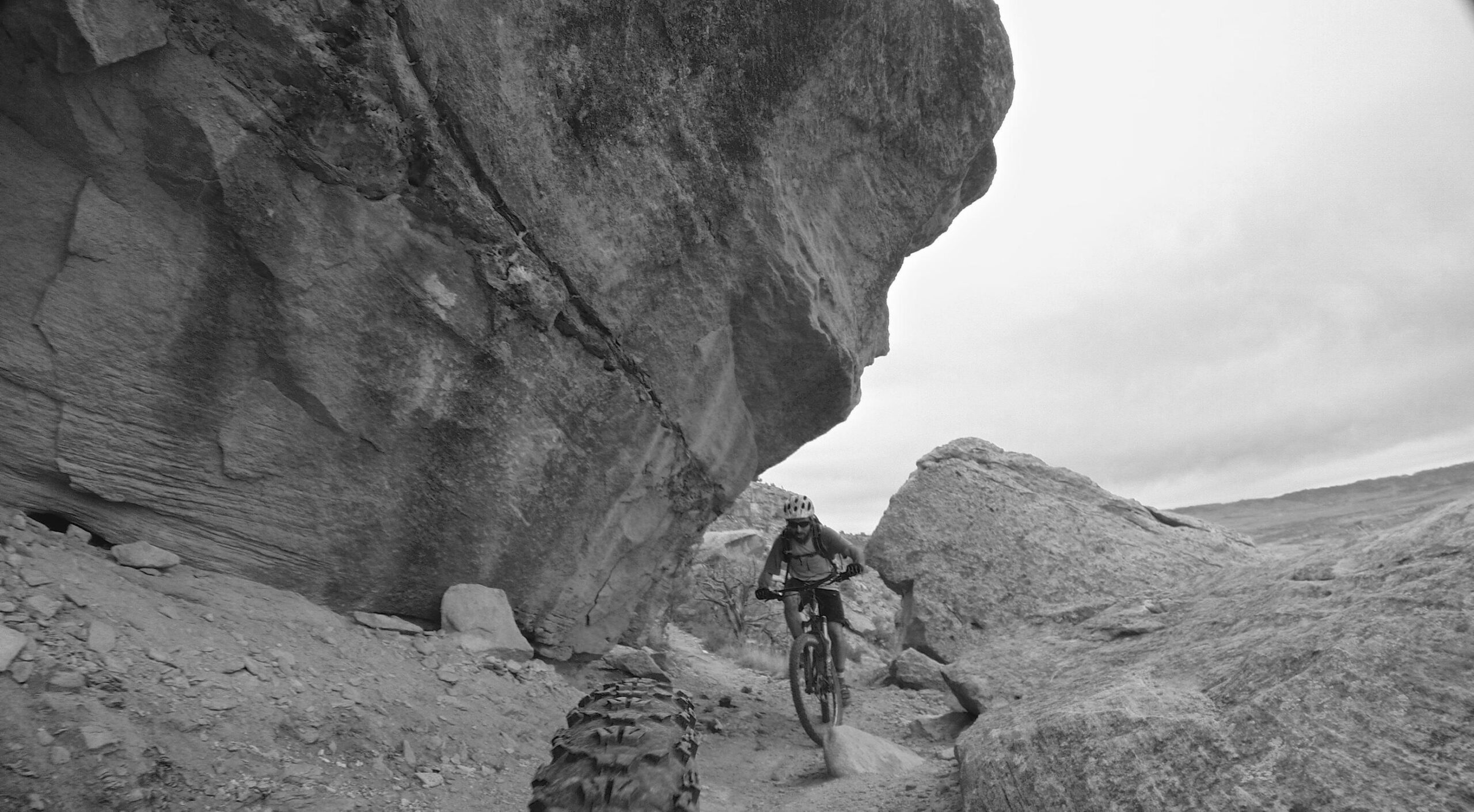 A black and white image of a mountain biker riding through a narrow rocky path surrounded by large boulders and stone formations. The biker is wearing a helmet and appears focused as they navigate the terrain. The sky is overcast, adding to the rugged outdoor setting. Kokopelli Area Trails mountain bike trail.