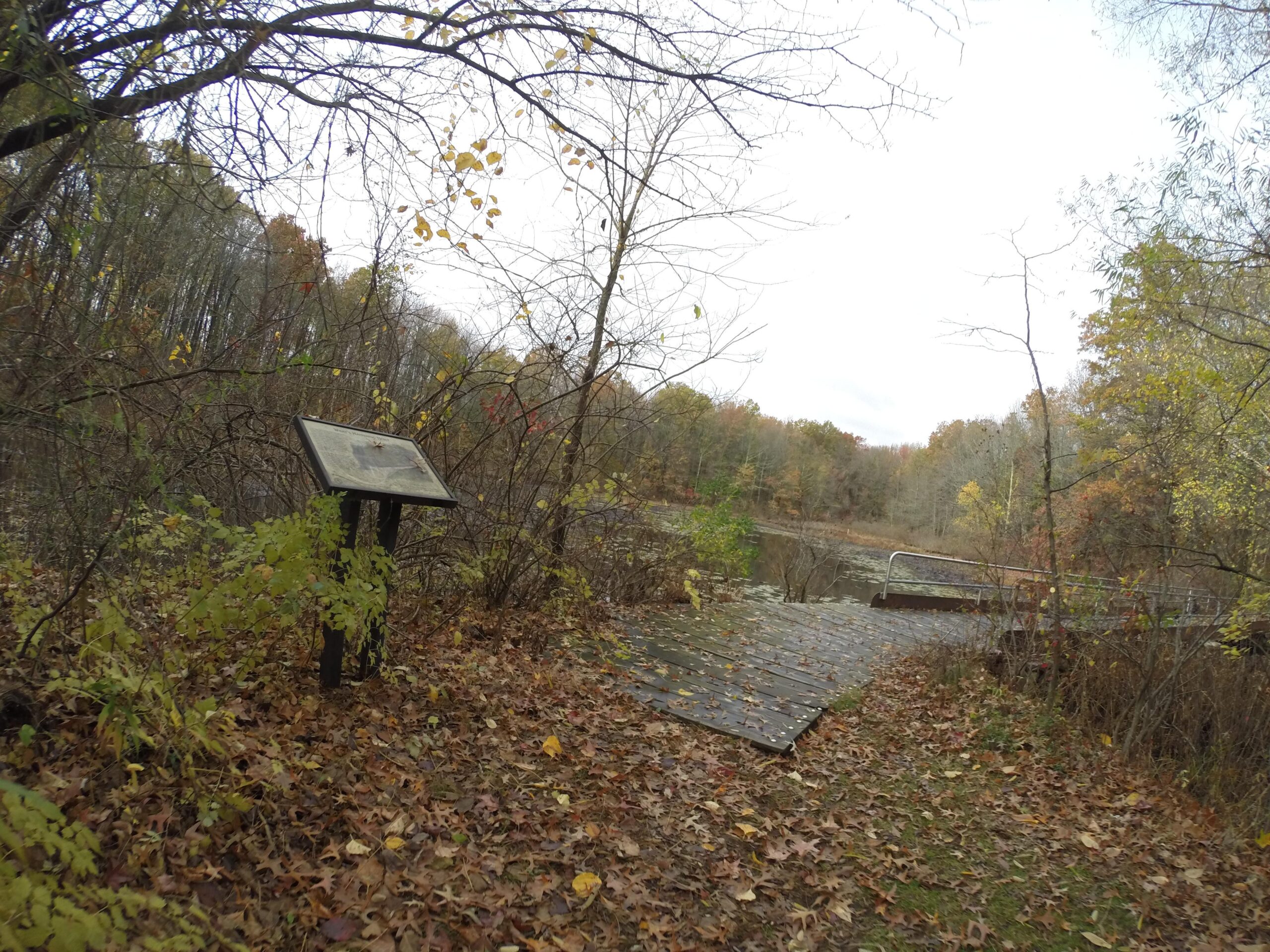 A scenic view of a wooded area in autumn, featuring a sign positioned among fallen leaves and shrubs. In the background, a small, serene body of water is visible, along with a wooden dock leading out over the water. The trees are displaying various fall colors, and the sky is overcast. Wolfes Pond park mountain bike trail.