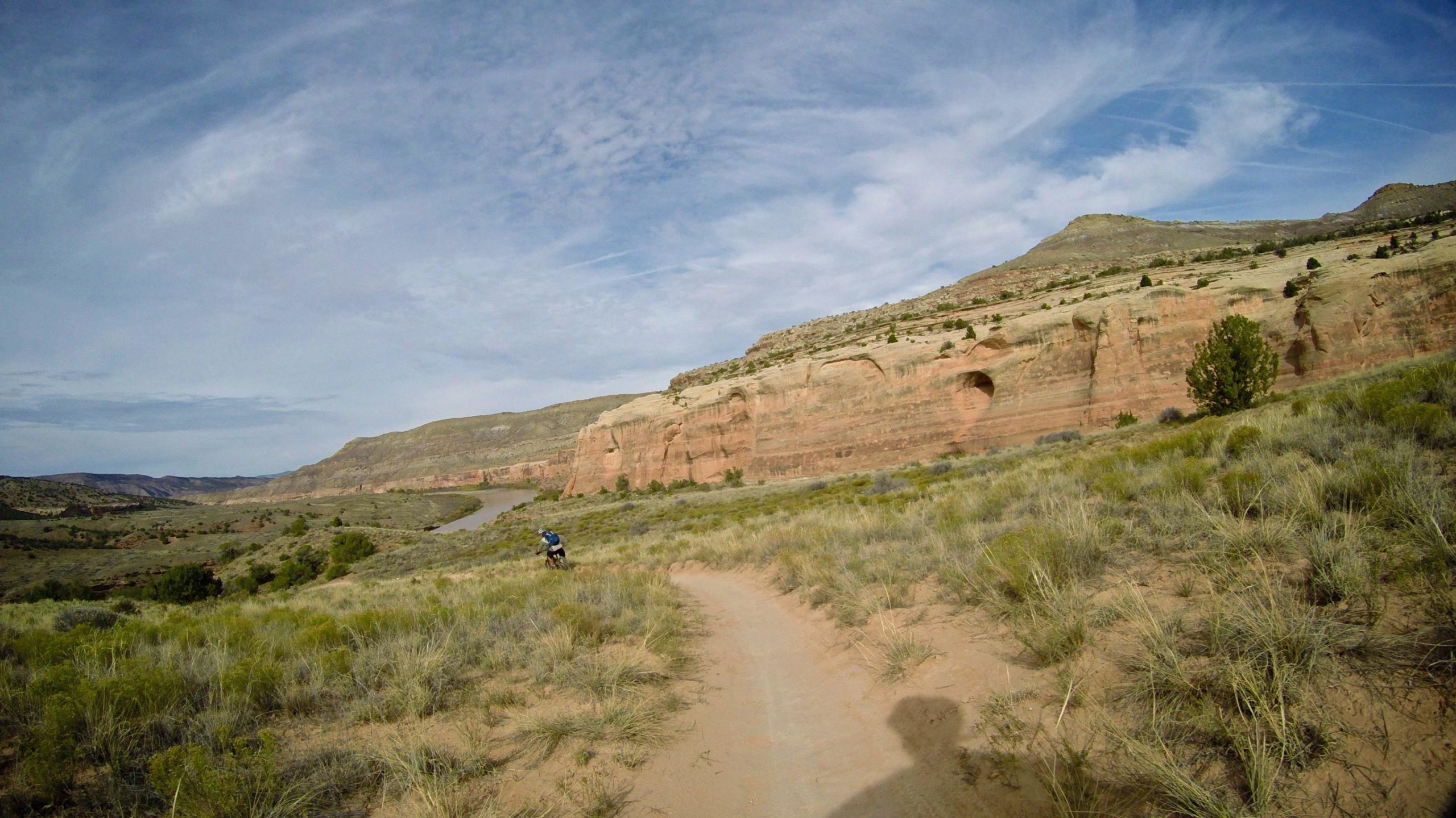 A scenic view of a winding dirt path through grassy terrain, surrounded by rocky cliffs and a river in the valley below. The sky is partly cloudy with visible blue patches, and a person on a bicycle is riding along the trail. Kokopelli Area Trails mountain bike trail.