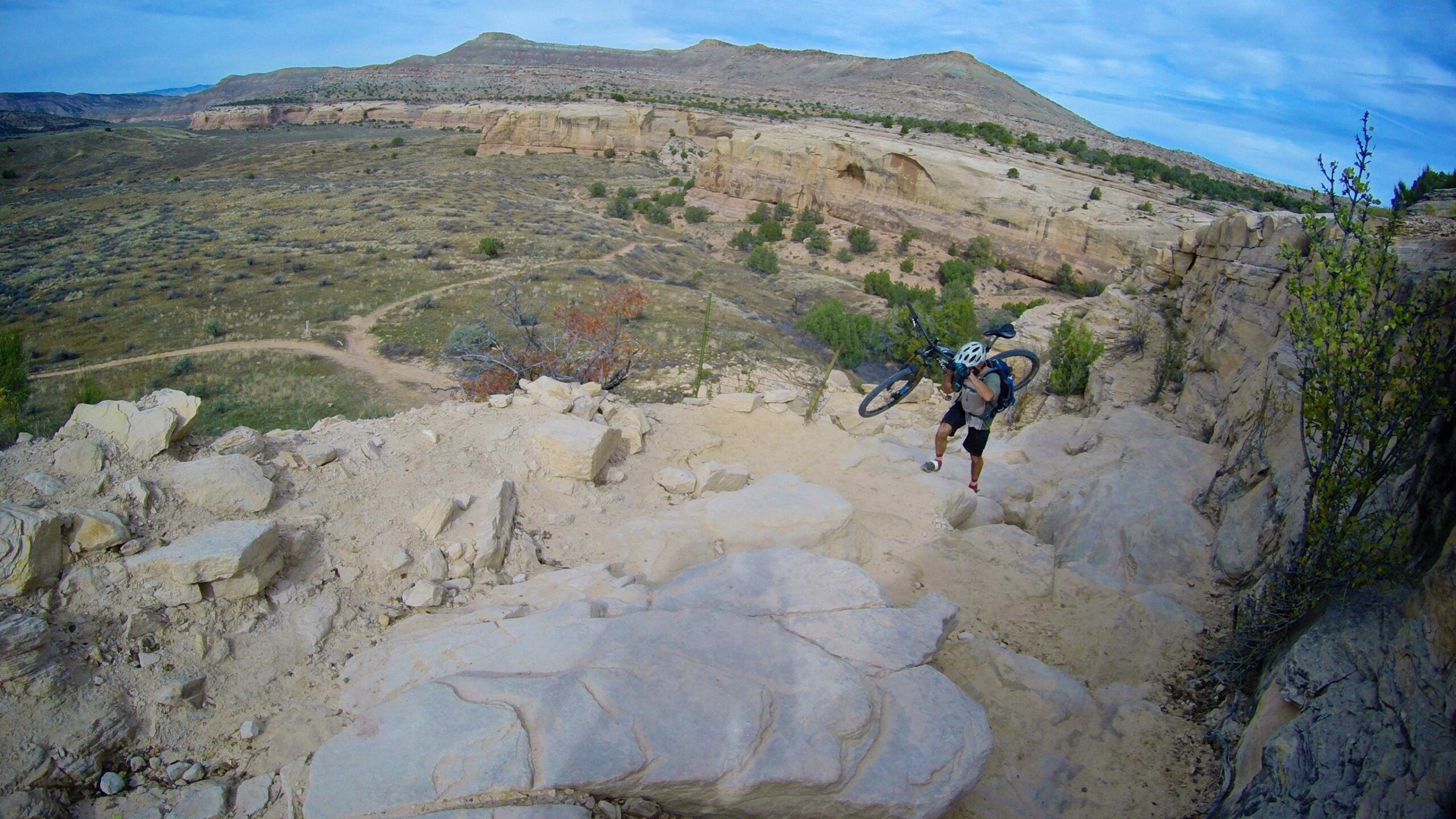 A mountain biker carrying their bike while navigating a rocky terrain on a hillside, with a panoramic view of a vast landscape featuring rolling hills and sparse vegetation under a partly cloudy sky. Kokopelli Area Trails mountain bike trail.