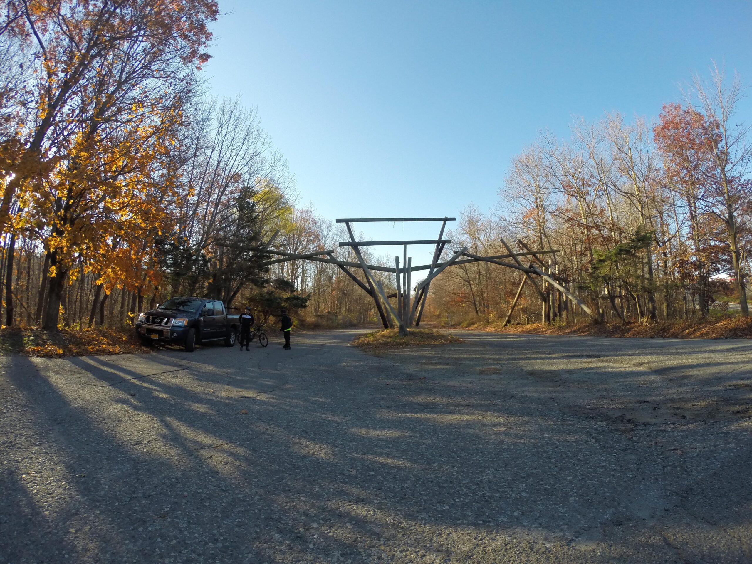 A gravel path in a wooded area during autumn, featuring a black vehicle parked on the left. Two individuals, one holding a bicycle, stand near the vehicle. In the background, there is a structure made of wooden beams, set against a clear blue sky with trees displaying fall foliage. Long Pond Ironworks State Park mountain bike trail.