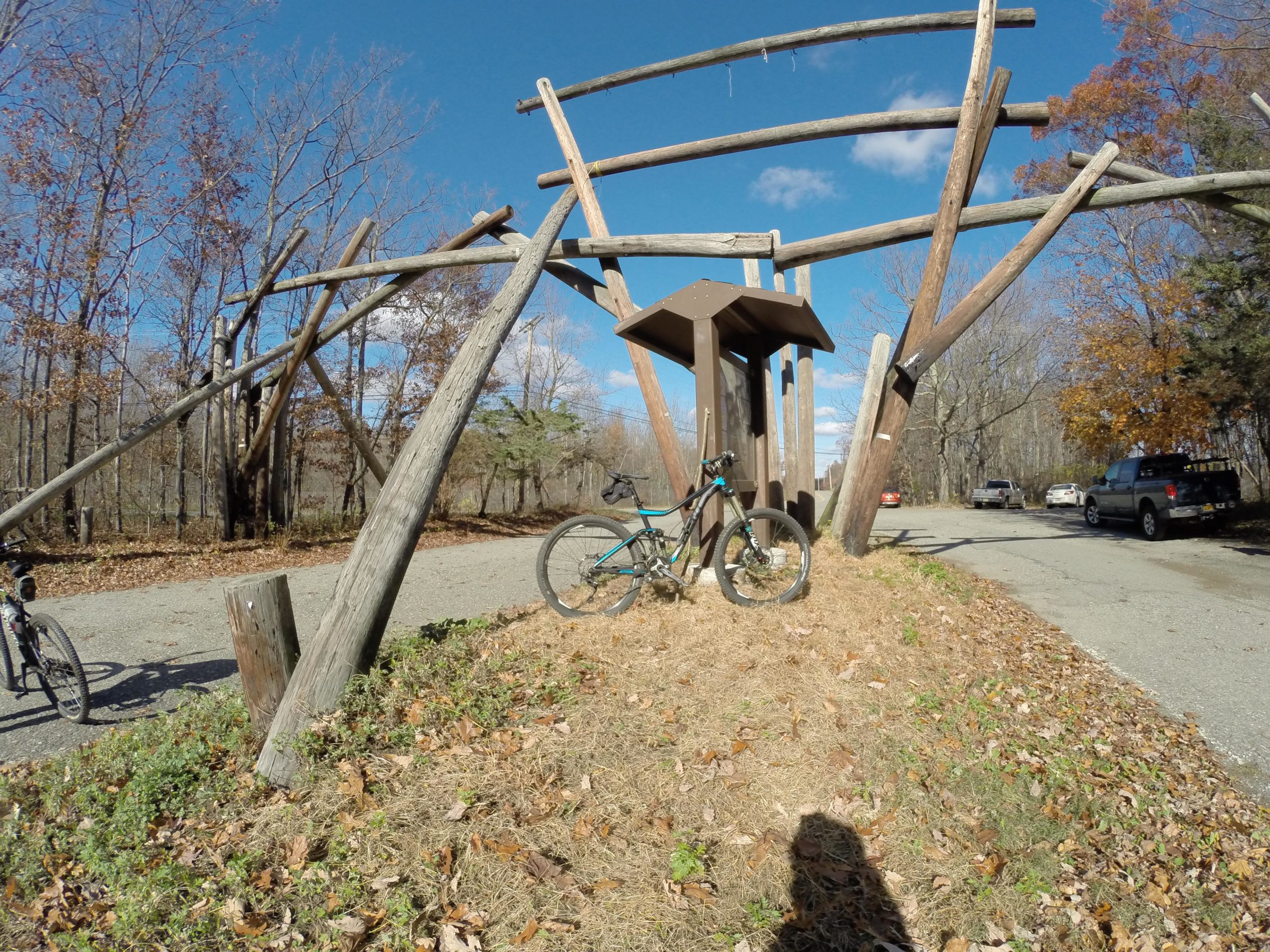 A mountain bike rests on the ground near a unique wooden structure made of large logs, set against a backdrop of trees with autumn leaves. A gravel path runs alongside the scene, with several cars visible in the background under a clear sky. Long Pond Ironworks State Park mountain bike trail.
