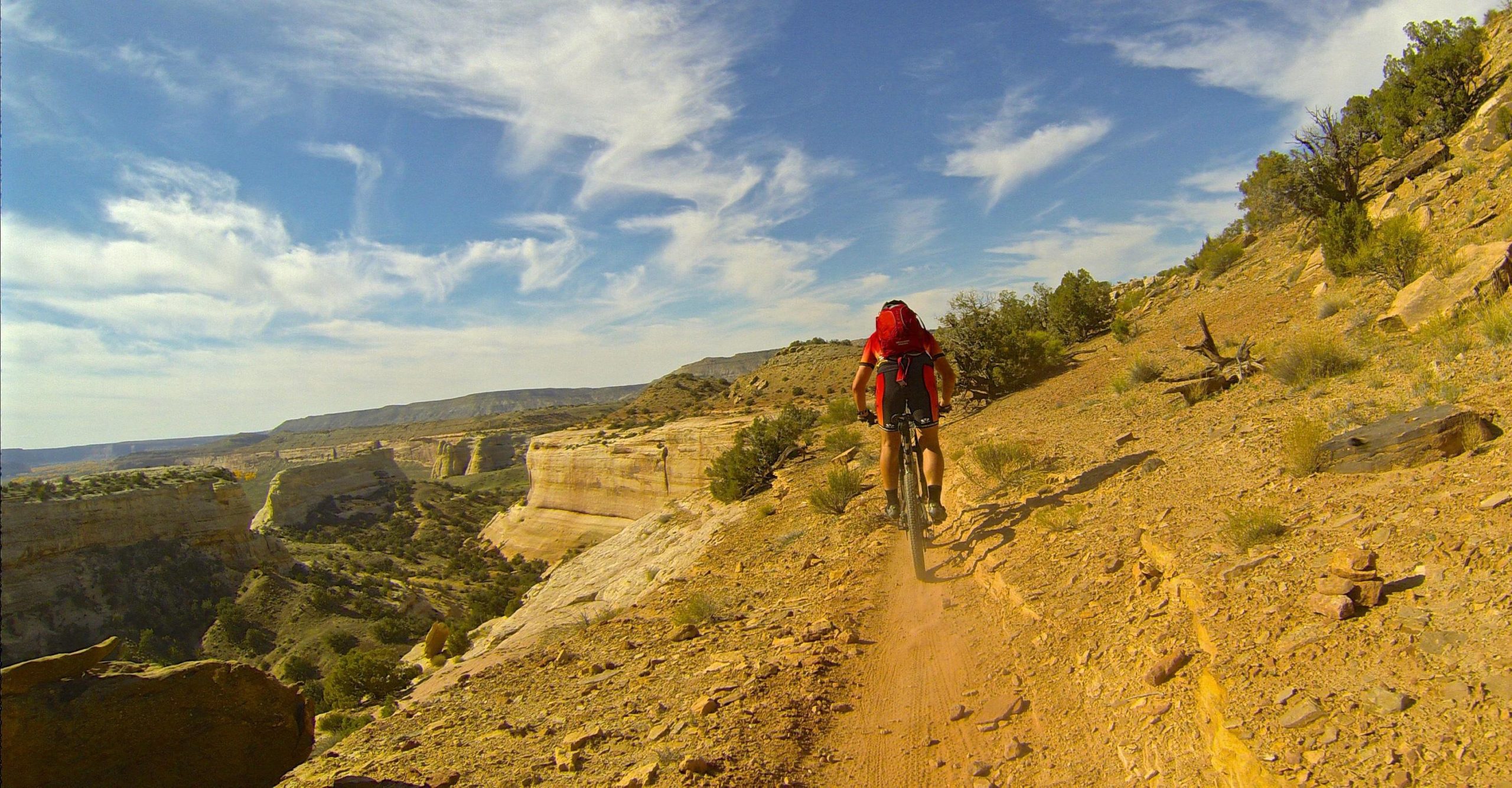 A person riding a mountain bike along a narrow trail through a rugged desert landscape with cliffs and sparse vegetation under a bright blue sky with wispy clouds. Western Rim mountain bike trail.