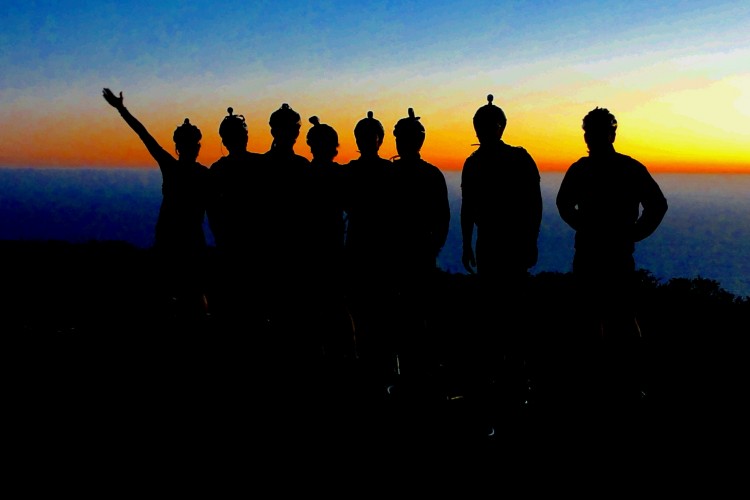 Silhouette of a group of seven people standing in front of a sunset, with the sun setting over the ocean. One person is raising an arm in celebration, while others stand side by side, all wearing hats that resemble small helmets. The sky transitions from blue to orange, creating a vibrant backdrop.