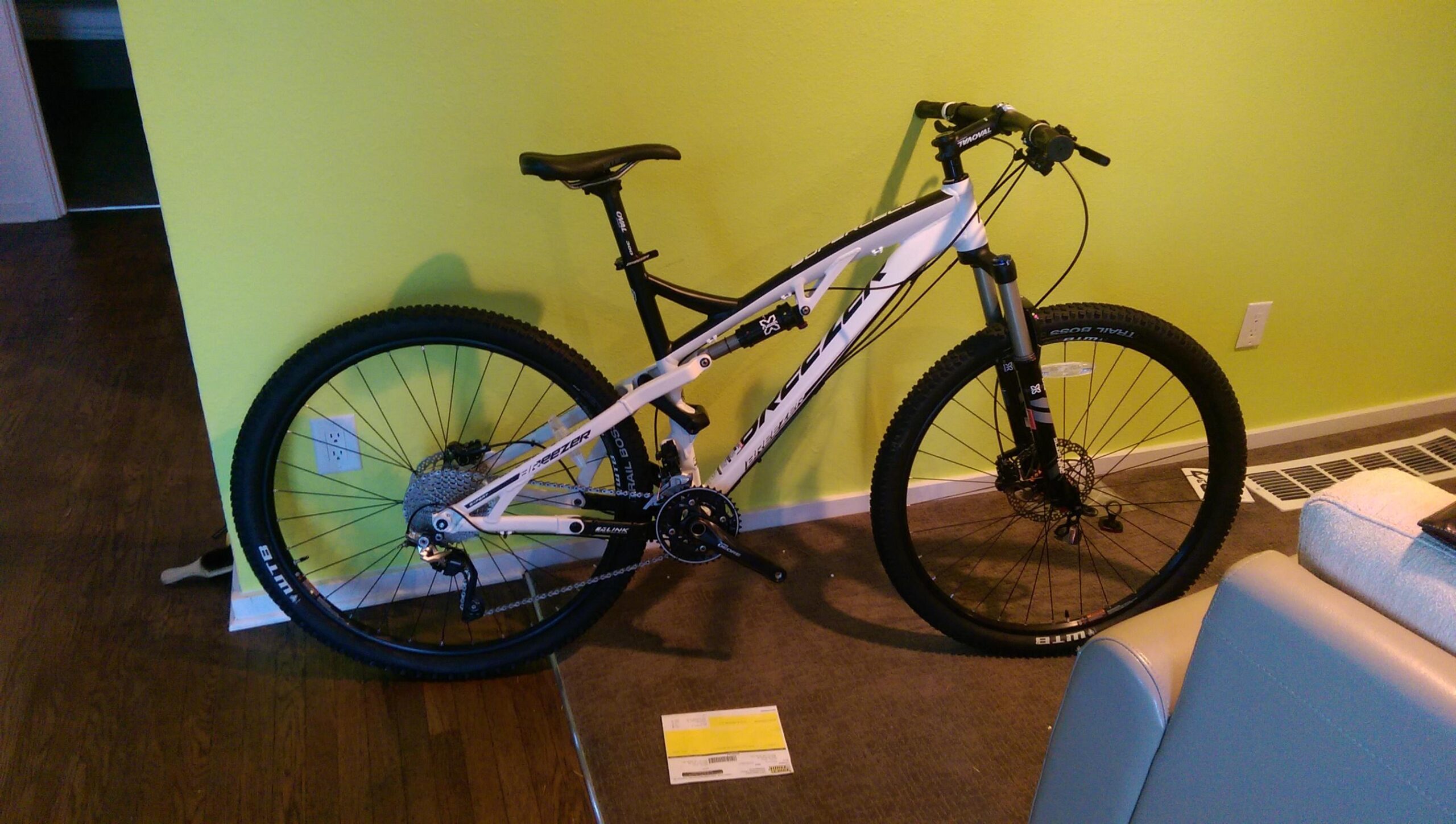 Breezer Supercell Pro: A white mountain bike positioned against a bright green wall, with visible details of its frame, wheels, and components. The bike is placed on a carpeted floor next to a light-colored couch, and a piece of mail is lying on the floor nearby.