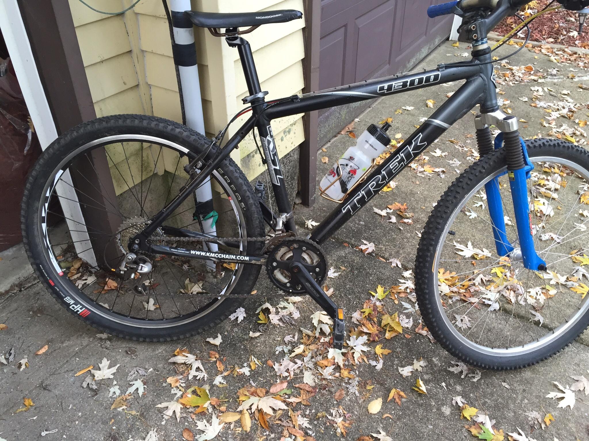 Trek 4300: A black Trek mountain bike resting against a garage wall, surrounded by fallen autumn leaves. The bike features knobby tires and a water bottle holder, with some dirt visible on the frame indicating recent use.