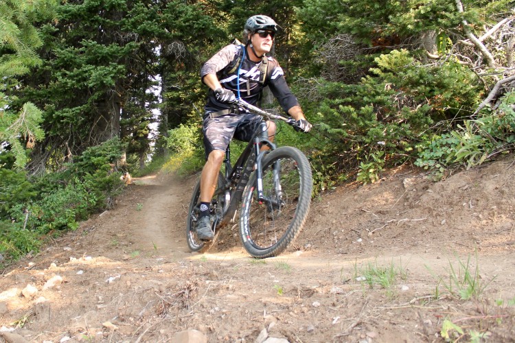 A mountain biker riding a bike on a dirt trail surrounded by trees and greenery. The cyclist is wearing a helmet, sunglasses, and a dark athletic outfit, navigating a curve on the trail.