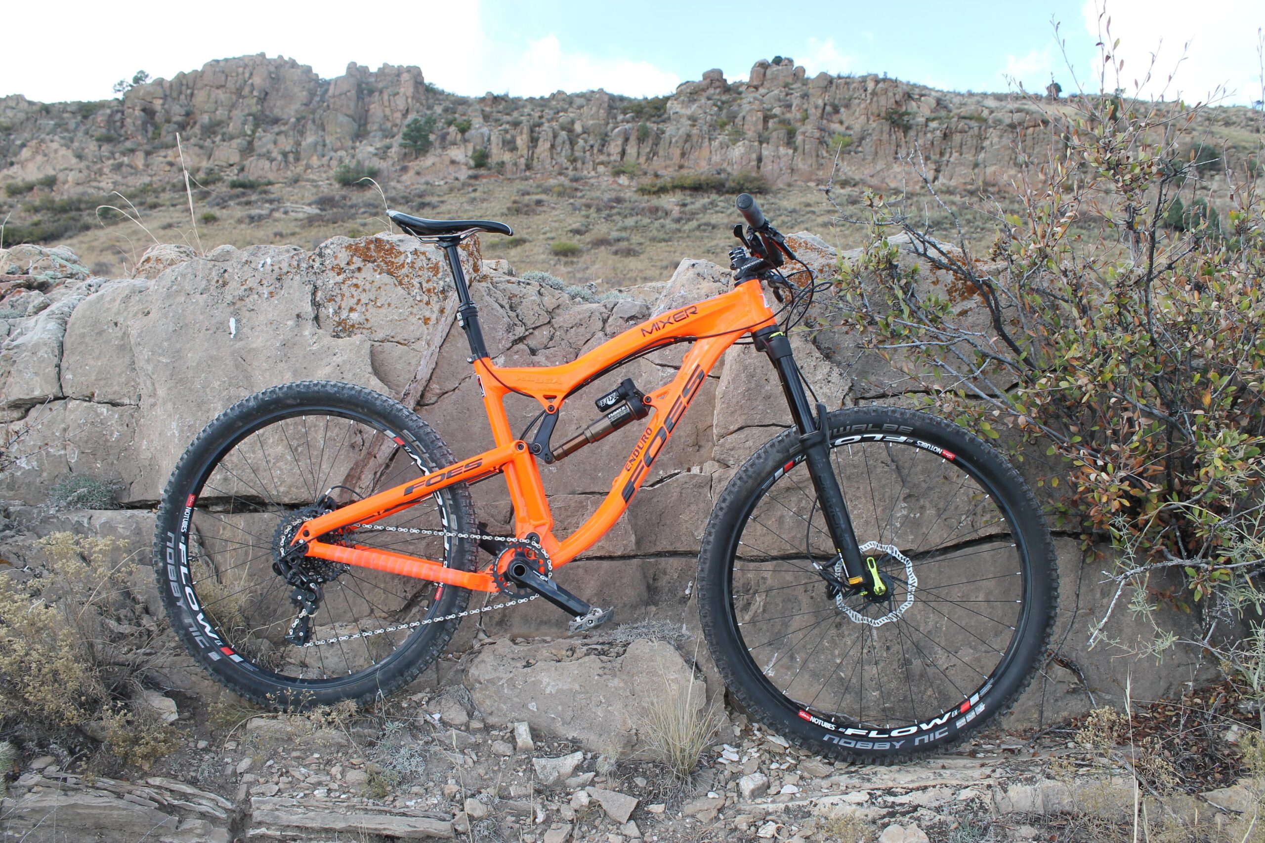 Foes Mixer Enduro: A vibrant orange mountain bike leaning against a rocky outcrop, surrounded by sparse vegetation and rugged terrain. The bike features large tires, a sleek frame, and a mountain bike saddle, set against a backdrop of rocky hills and a blue sky.