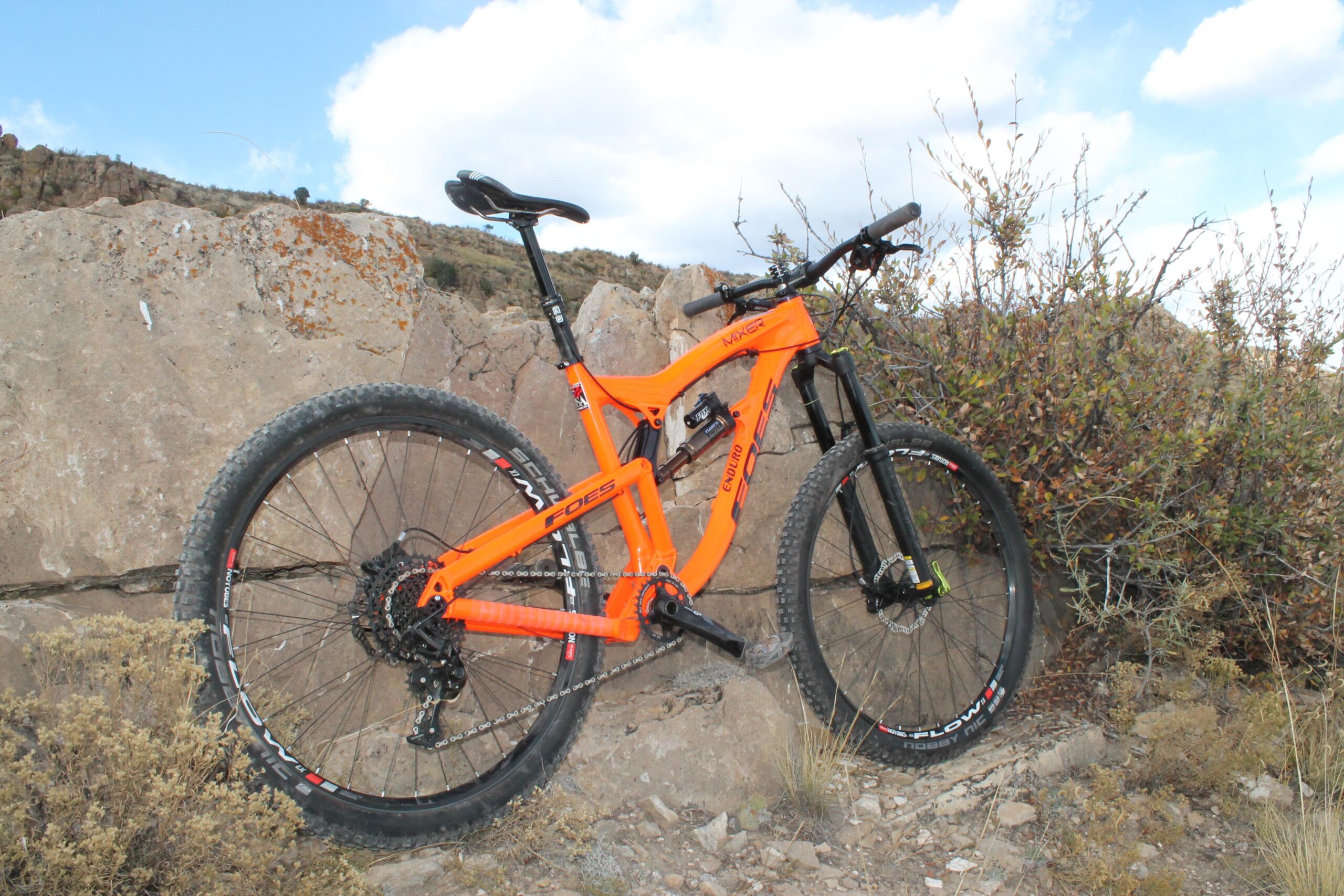 Foes Mixer Enduro: An orange mountain bike leaning against a large rock, surrounded by dry grass and shrubs under a partly cloudy sky. The bike features a full suspension design and robust tires suitable for off-road terrain.