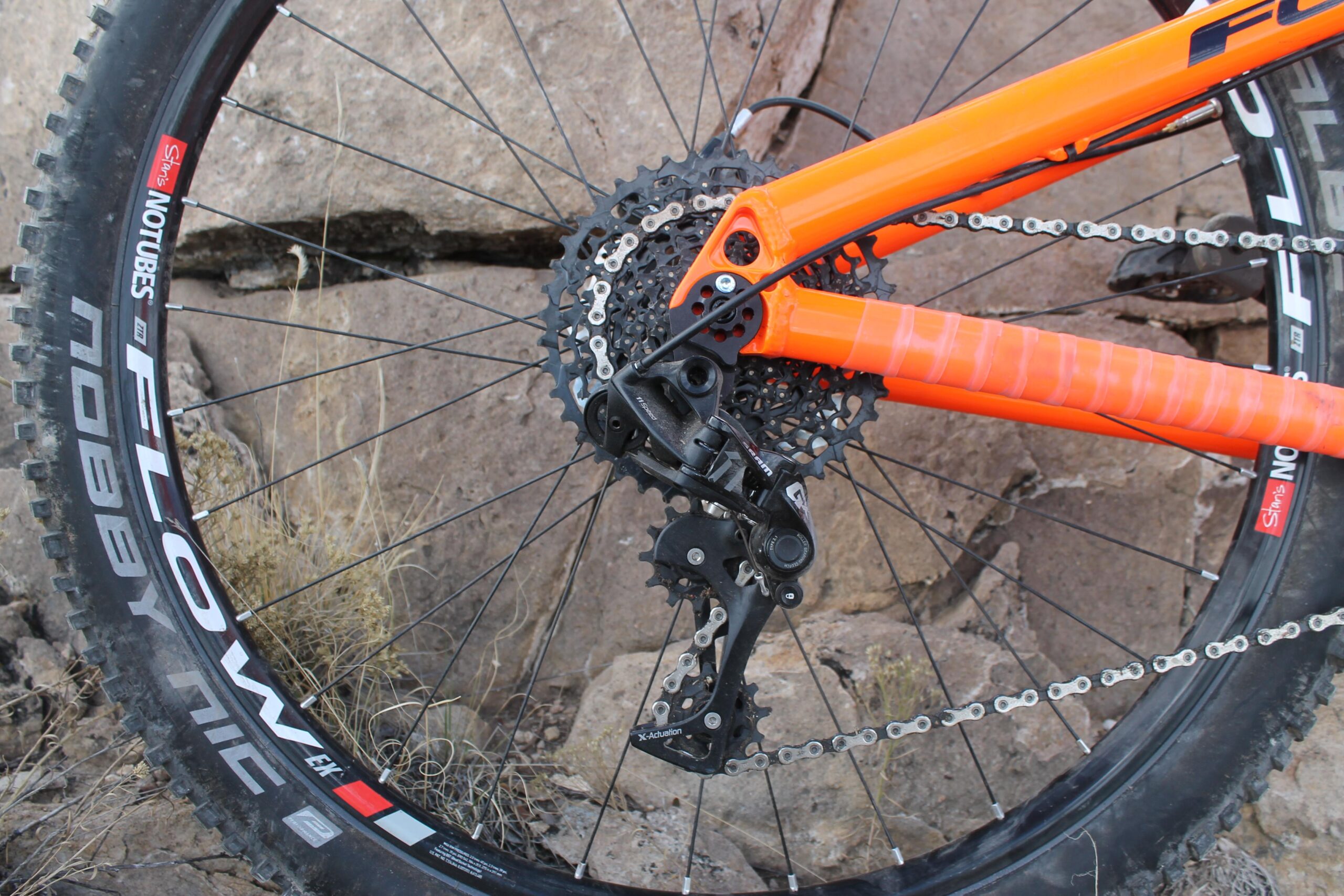 Foes Mixer Enduro: Close-up view of the rear wheel and drivetrain of a mountain bike, featuring an orange frame, black derailleur, and a spoked wheel with a "Flow" tire on rocky terrain.