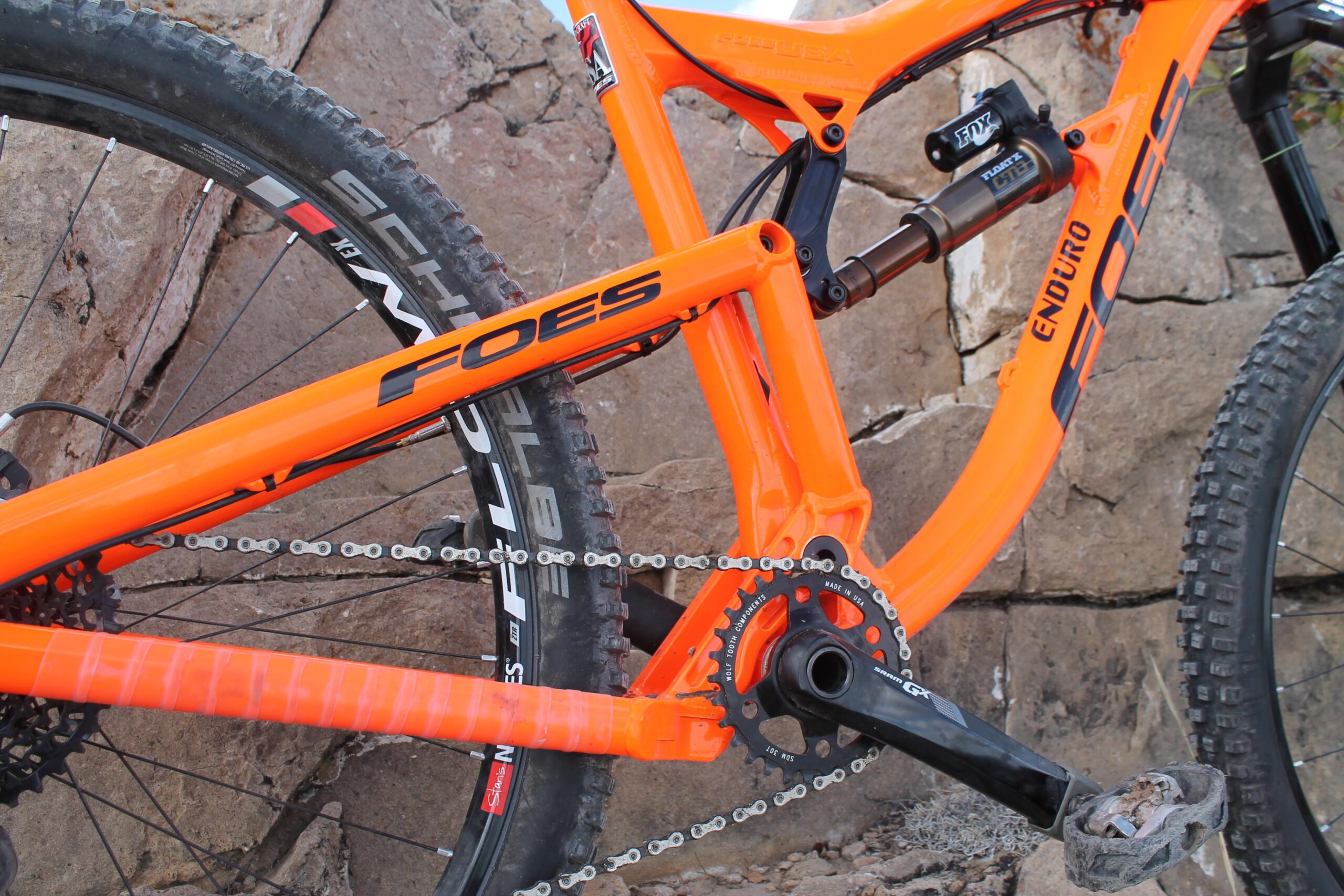 Foes Mixer Enduro: Close-up view of a Foes mountain bike frame in bright orange, showcasing the chainring, pedals, and rear suspension components, with textured tires and rocky terrain in the background.