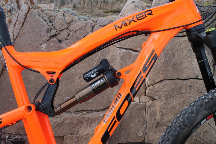Close-up view of the frame of an orange mountain bike, featuring the model name "FOES MIXER" prominently displayed. The bike is equipped with a FOX shock absorber for suspension. The background consists of rocky terrain, indicating an outdoor setting suitable for off-road cycling.
