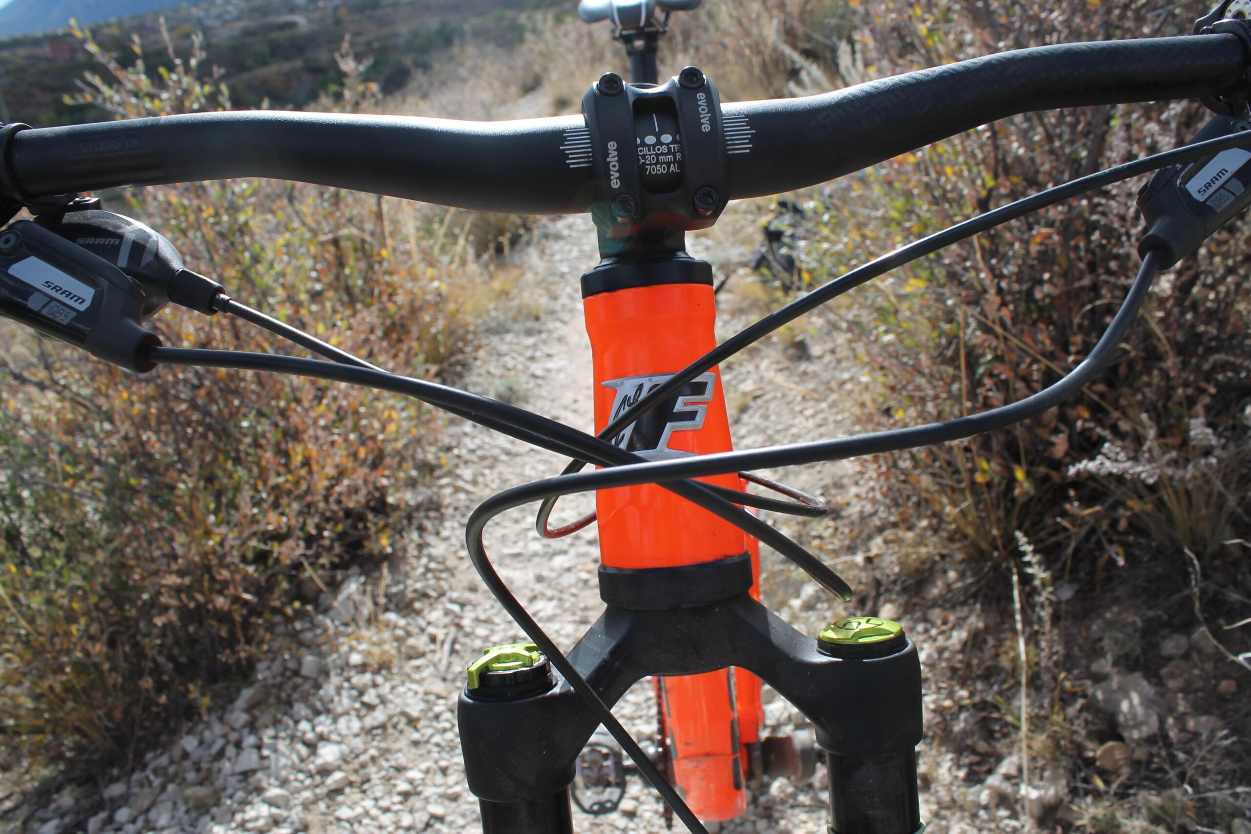 Foes Mixer Enduro: Close-up view of a mountain bike's handlebars and front fork, featuring a vibrant orange frame, SRAM shifters, and black cables set against a rocky trail with surrounding vegetation.