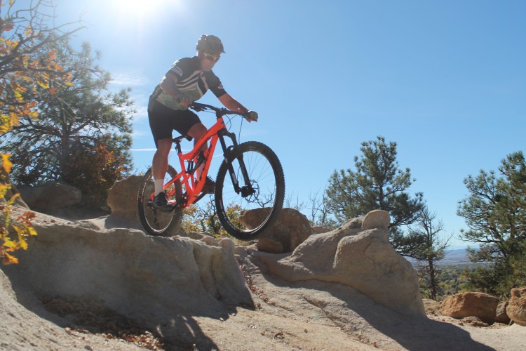 A person riding a bright orange mountain bike over rocky terrain, with trees and a clear blue sky in the background. The cyclist is wearing a helmet and sports attire while navigating the rocky trail in a sunlit outdoor setting.