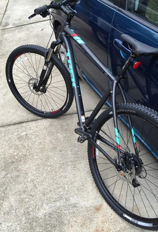 Trek X-Caliber 7: A close-up view of a black mountain bike with teal and red accents, positioned next to a blue car on a concrete surface. The bike features thick tires and front suspension, suitable for off-road riding.