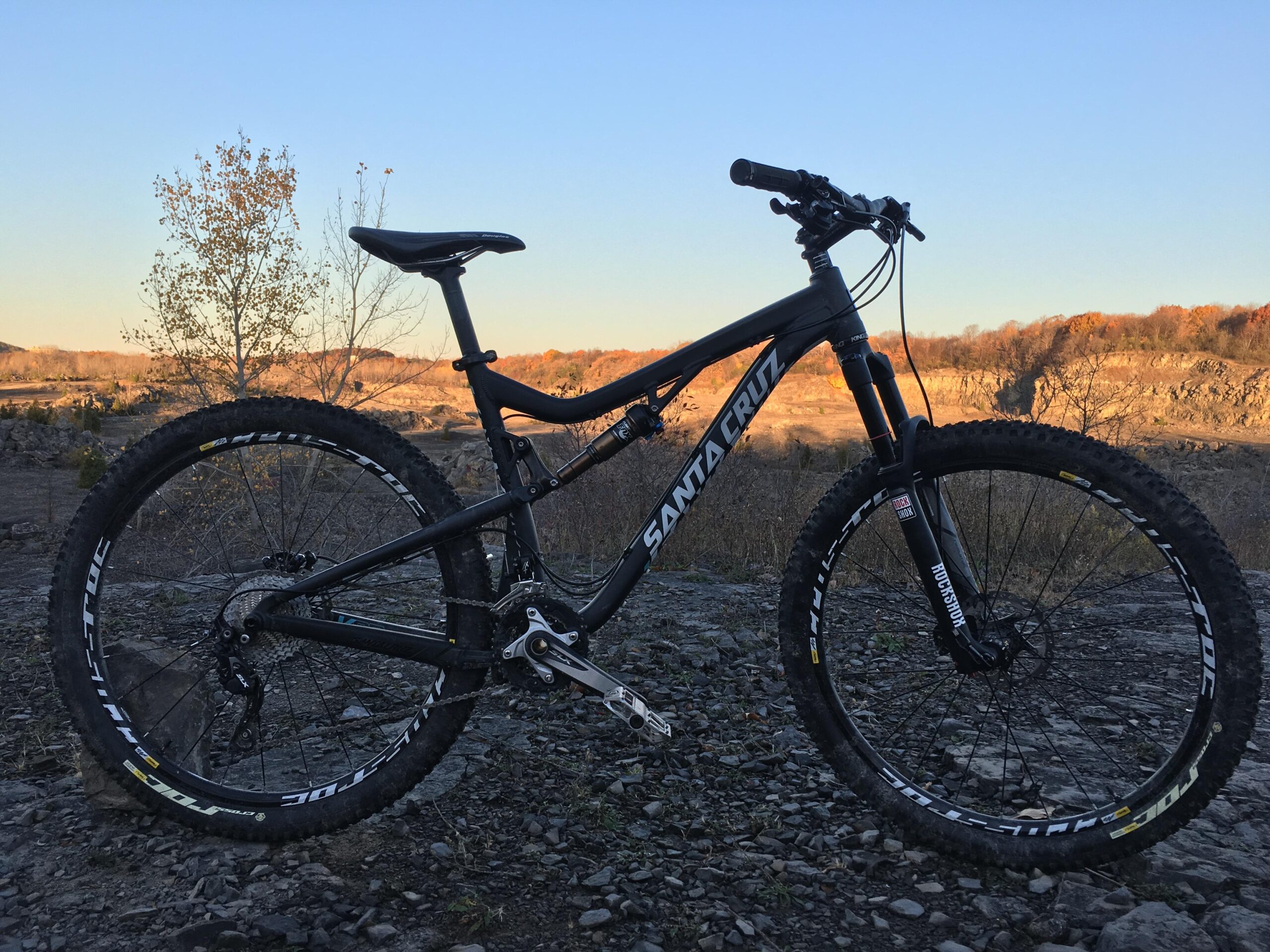 Santa Cruz 5010: A black mountain bike with "Santa Cruz" branding is positioned on rocky terrain, with a backdrop of a sunset illuminating the landscape. Bare trees and fall foliage are visible in the background, suggesting an outdoor setting suitable for biking.