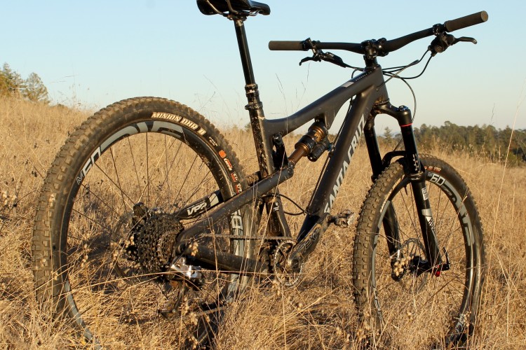 A black mountain bike resting in tall, dry grass under a clear sky, showcasing its rugged tires and sleek frame. The bike features prominent branding and is positioned at an angle, highlighting its suspension system and handlebars.