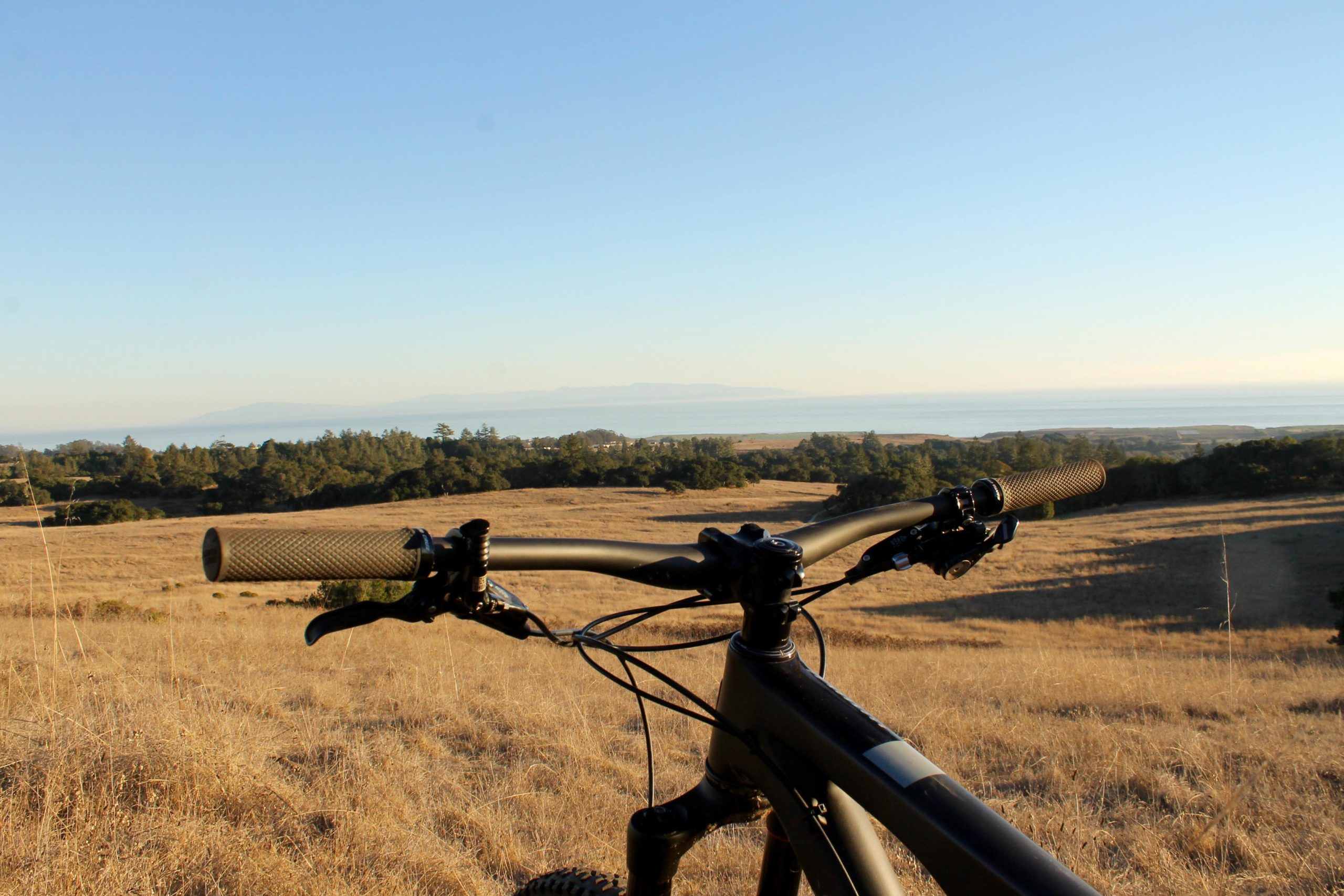 Close-up view of a mountain bike's handlebars overlooking a vast landscape with rolling hills and the ocean in the distance. The scene is bathed in warm golden light, indicating either sunrise or sunset, with a clear blue sky above. The foreground features golden grass, adding to the natural beauty of the setting. Wilder Ranch State Park mountain bike trail.