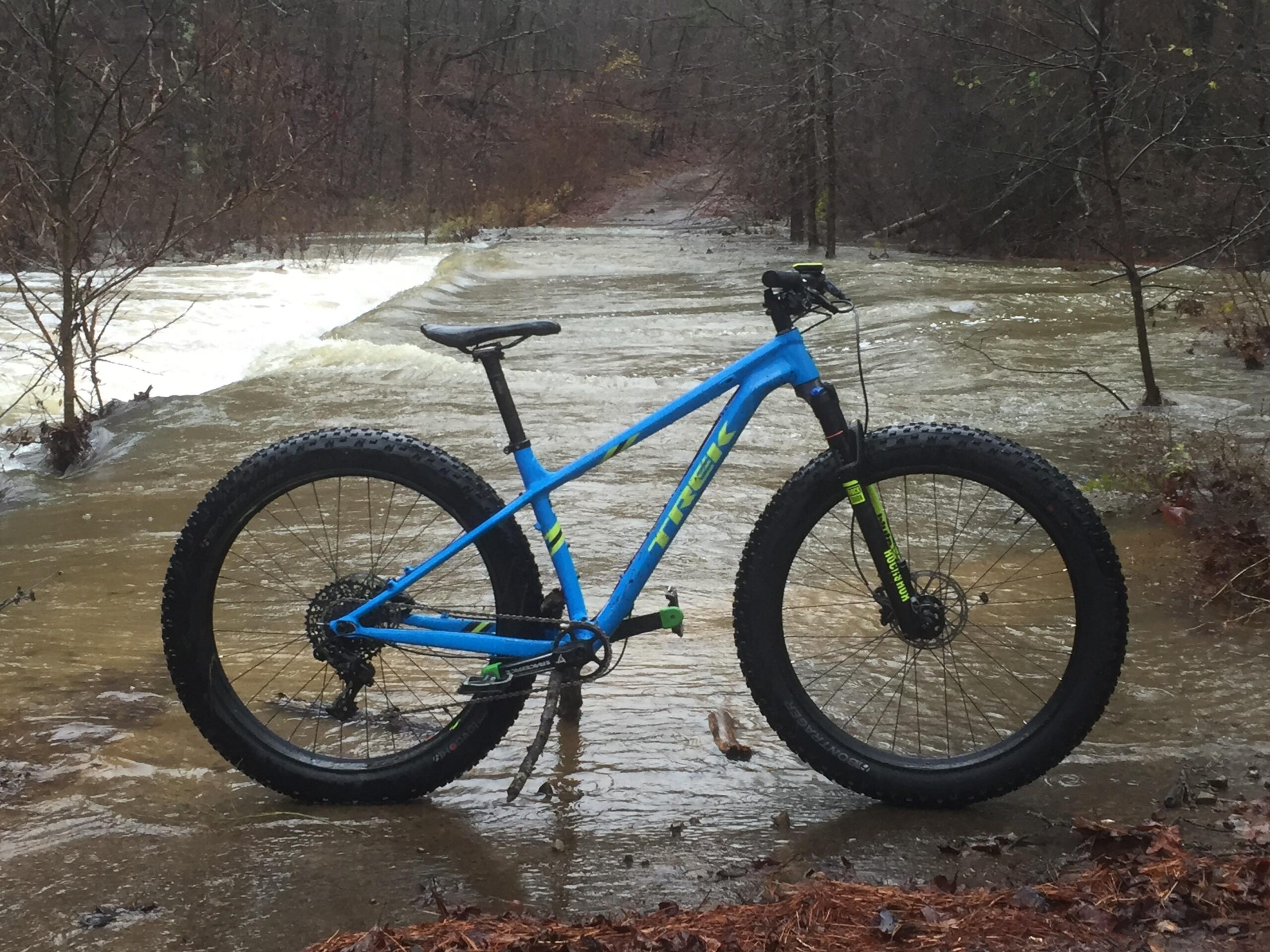 Trek Farley 9: A blue mountain bike is positioned next to a flooded trail, with water flowing over the path. Surrounding the scene are bare trees and muddy ground, indicating a rainy weather condition. The bike features wide tires suitable for off-road terrain.