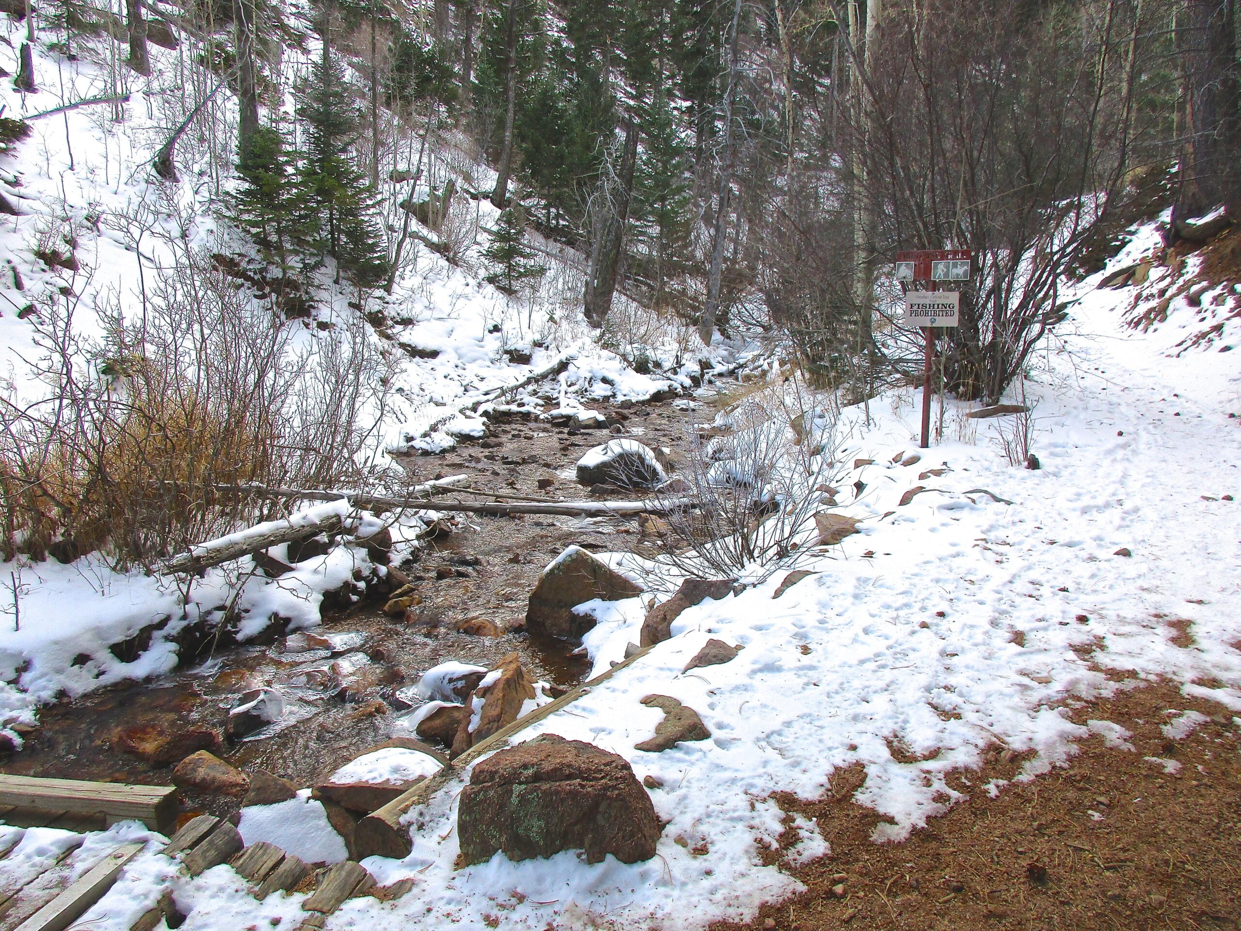A serene winter landscape featuring a snow-covered path alongside a flowing creek. The area is surrounded by trees and shrubs, with a sign indicating "Fishing Prohibited" visible near the bank of the stream. Snow blankets the ground, and logs are laid across the water, creating a natural, peaceful setting. Bear Creek Canyon Loop mountain bike trail.