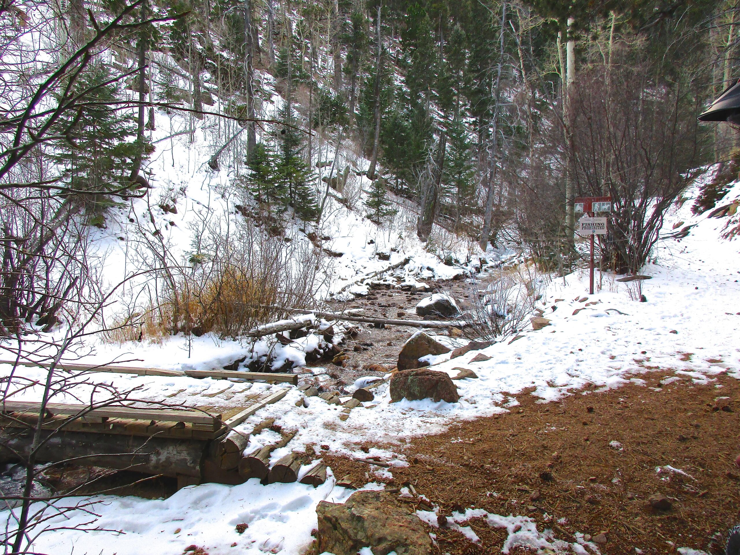 A serene winter landscape featuring a snow-covered creek bordered by evergreen trees and bare shrubs. A wooden footbridge crosses over the creek, which flows gently through a rocky bed. A sign indicating "Fishing Access" is visible along the shoreline, suggesting a recreational area in a forested setting. Bear Creek Canyon Loop mountain bike trail.